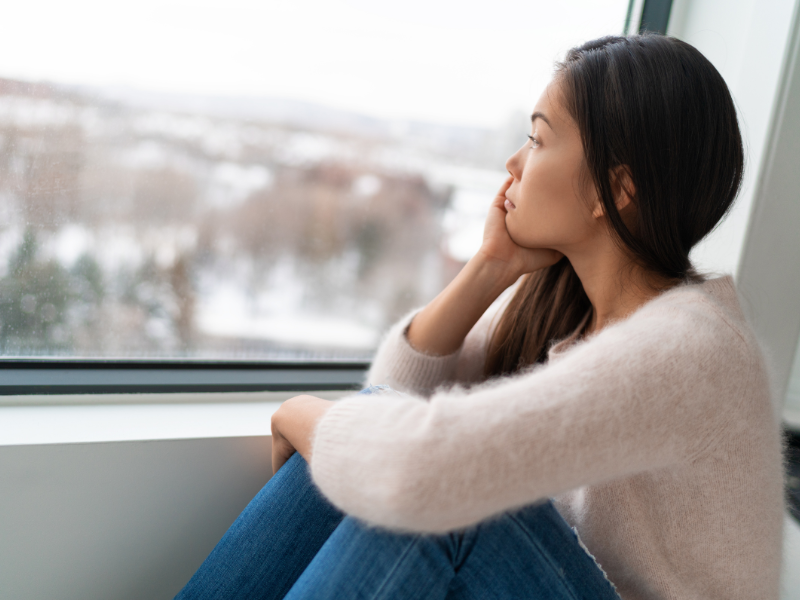 Woman in beige sweater sitting by window looking thoughtfully outside at winter landscape