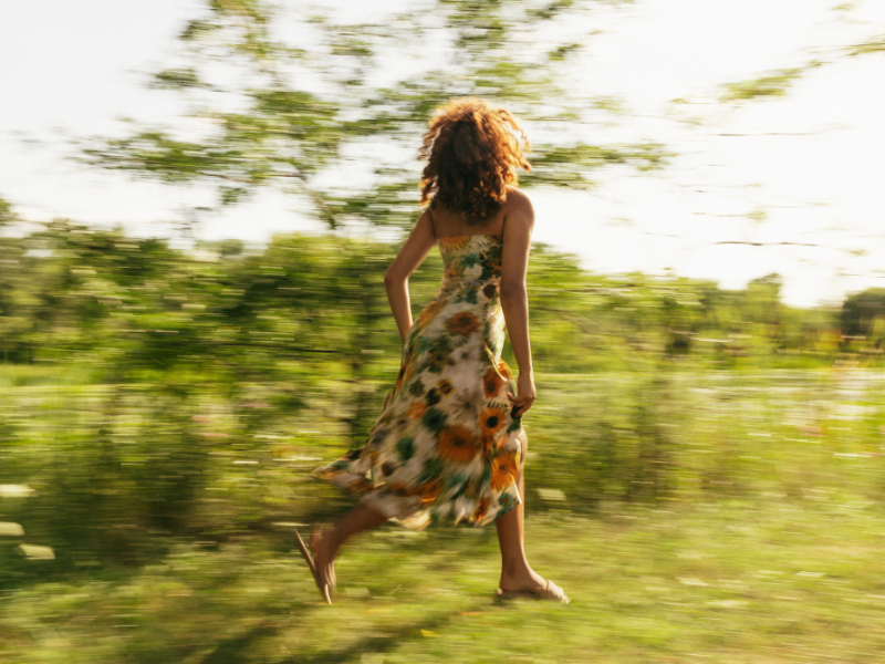Woman in flowing tie-dye dress running barefoot through green field with motion blur