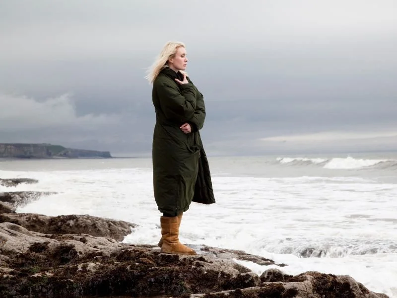 Woman in dark green coat standing on rocky coastline looking out at stormy ocean waves