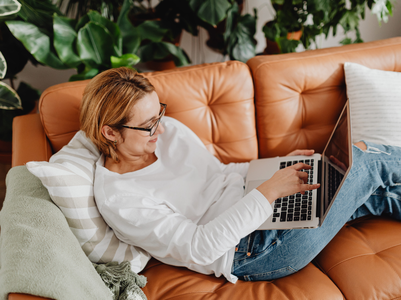 Woman in white sweater and glasses working on laptop while relaxing on tan leather couch with plants