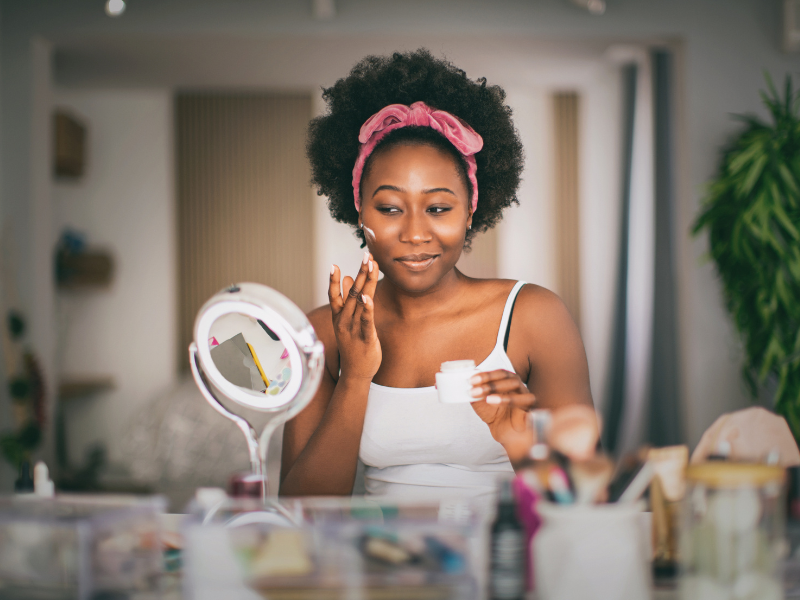 Woman with pink headband applying skincare product while looking in lighted mirror at vanity table