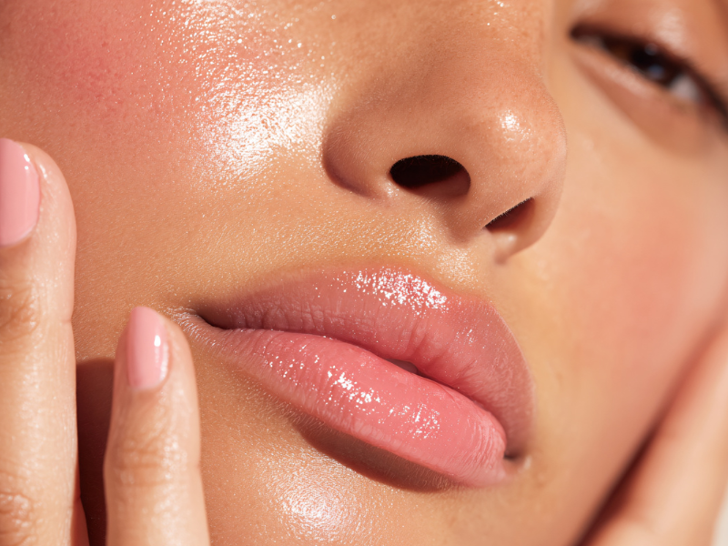 Close-up of woman's face showing dewy skin, glossy lips, and natural makeup with hand touching cheek