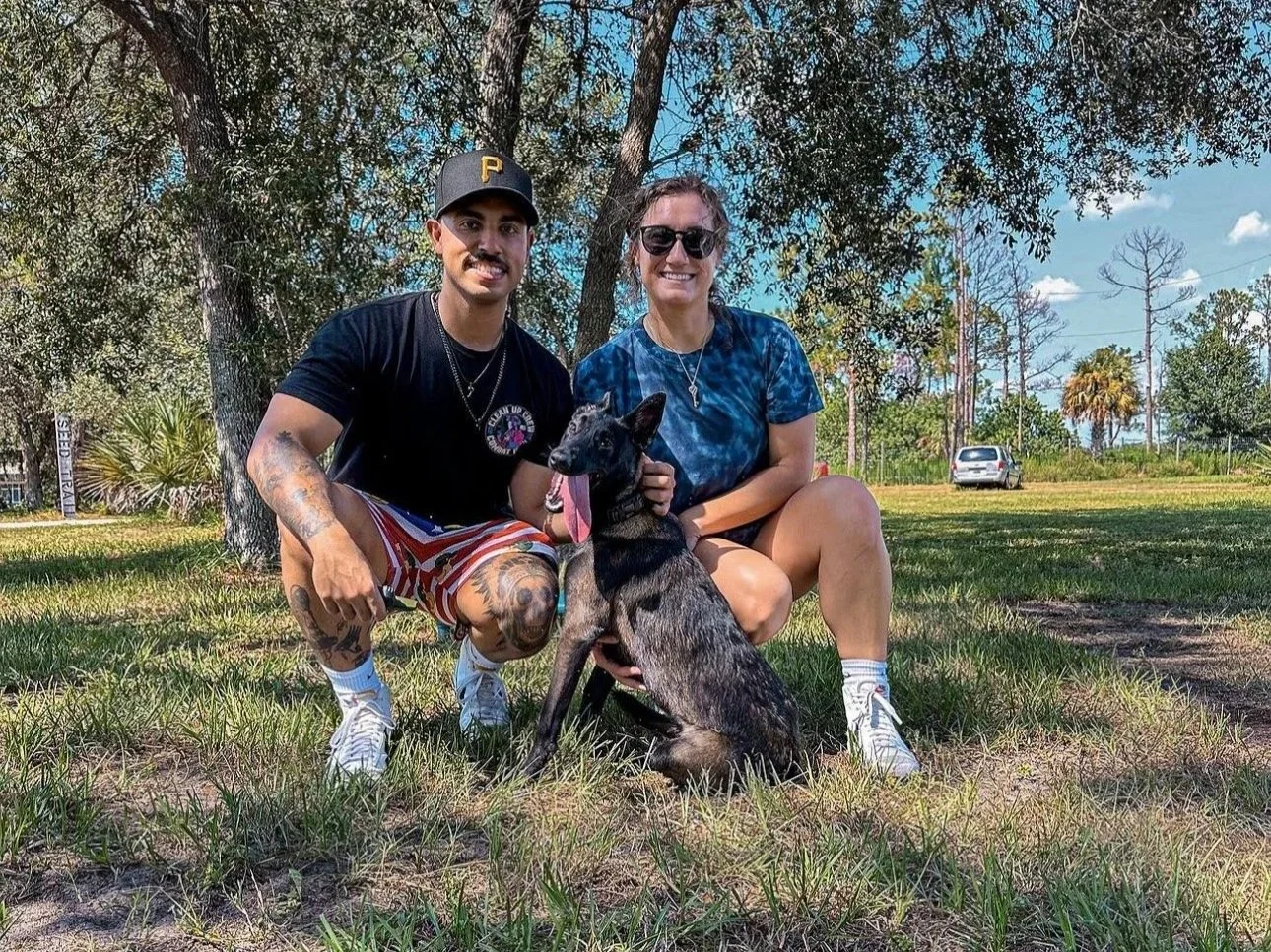 Two people sitting on grass under a tree with a dog, in a park with trees and a few cars in the background during the daytime.