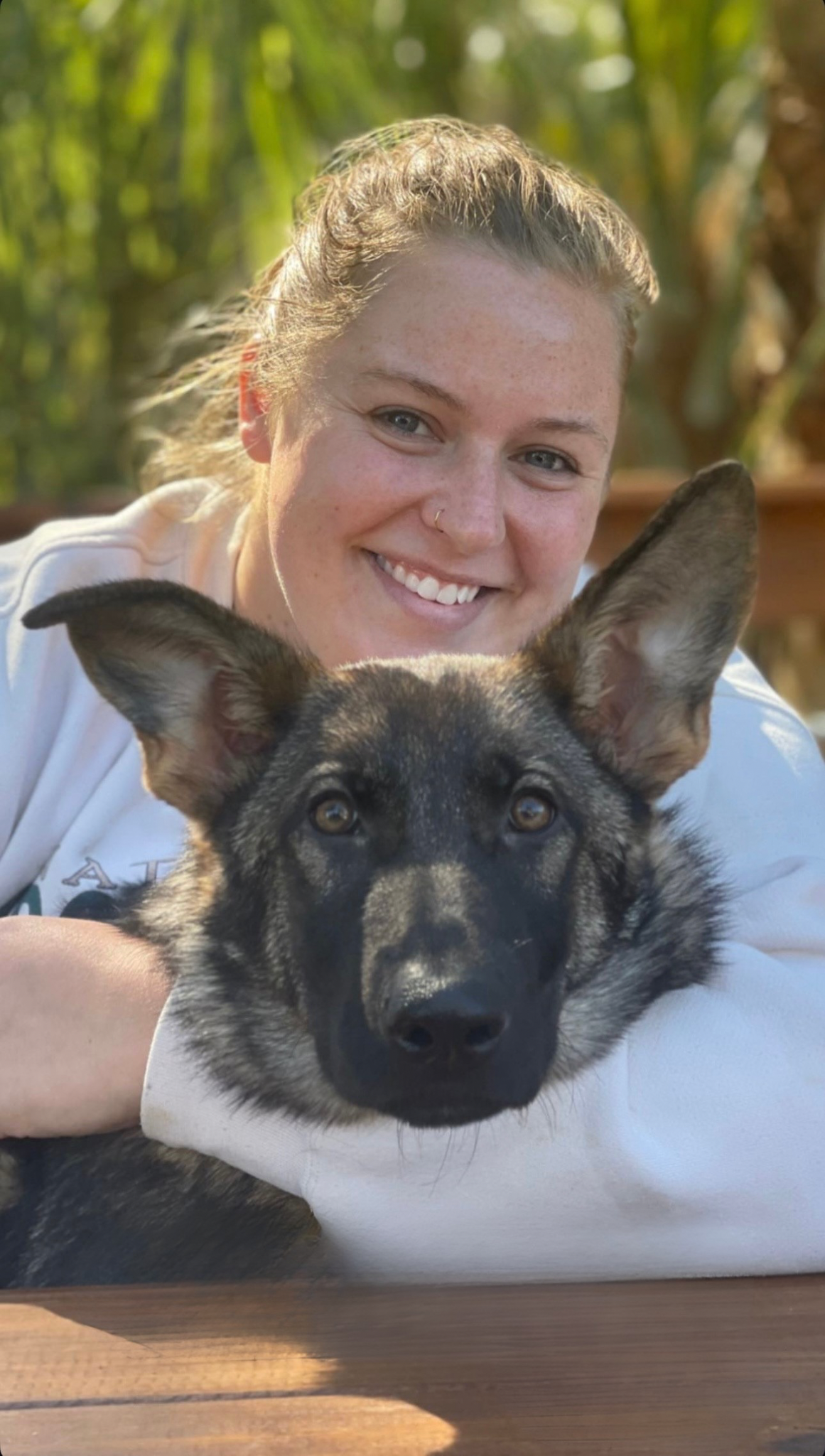 A woman with light skin and curly hair smiling while holding a black and tan dog with large ears in an outdoor setting with greenery.