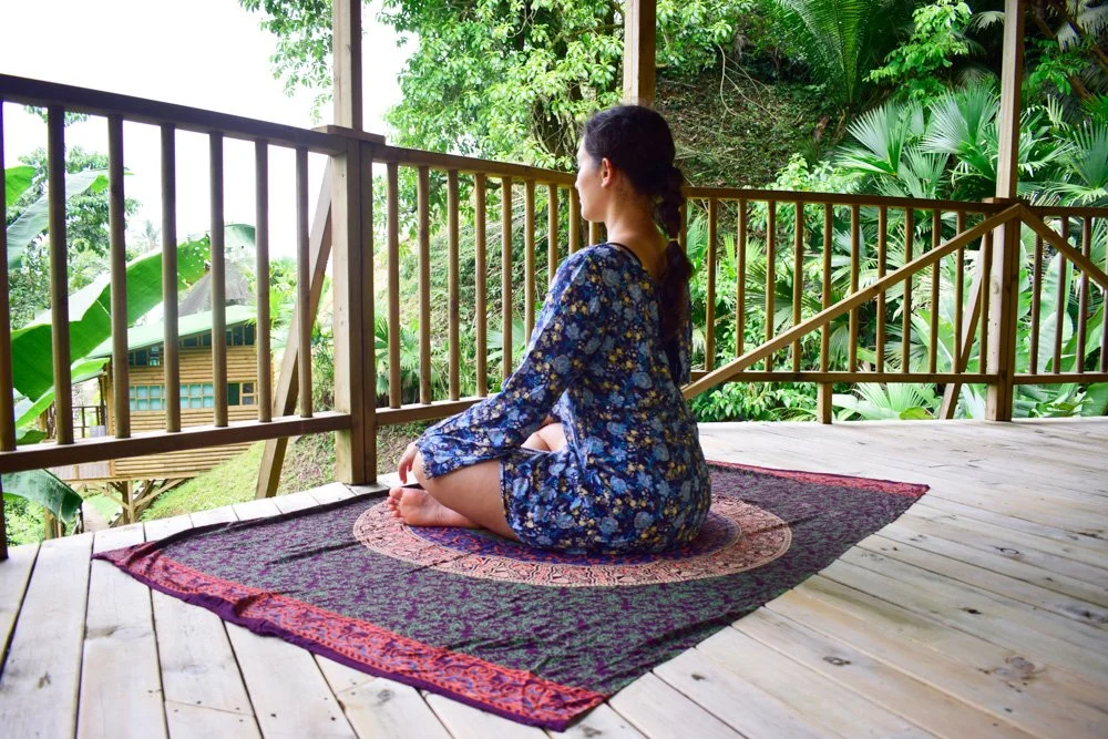 A woman in a blue floral dress practicing yoga or meditation on a rug on a wooden deck surrounded by lush green tropical plants.