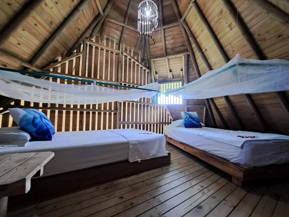Interior of a wooden A-frame cabin bedroom with two twin beds, mosquito nets hanging above, pillows with nautical designs, and a small balcony at the far end.