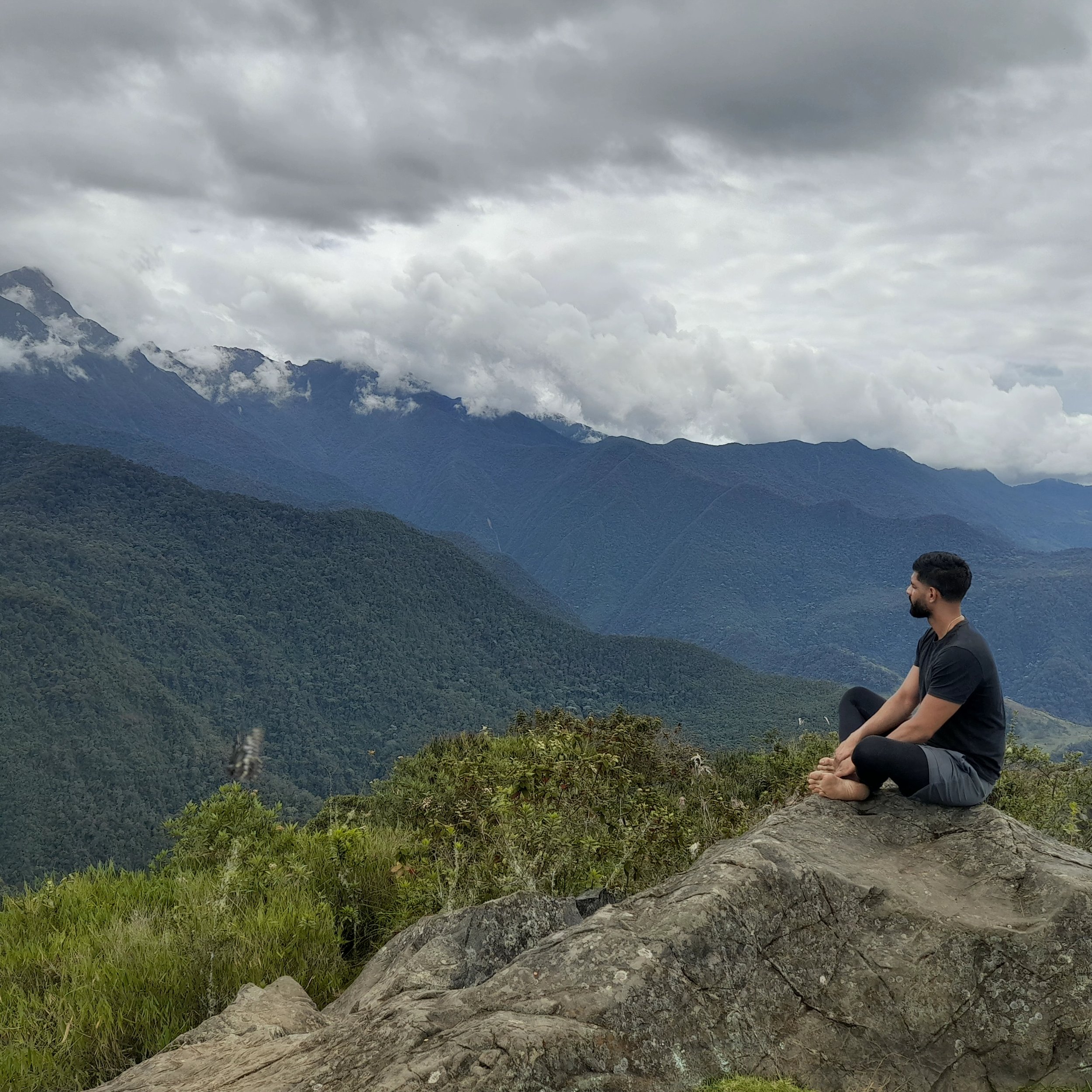 A man sitting on a rock in a mountainous landscape, looking at the mountains, with clouds overhead.
