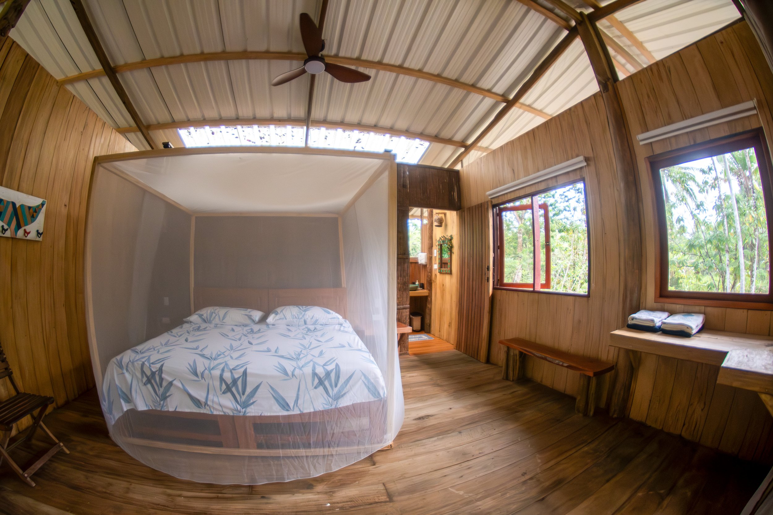 A cozy wooden cabin bedroom with three large open windows, a bed covered with a white and blue patterned duvet and mosquito net, a ceiling fan, a bench, towels, and a view of lush greenery outside.