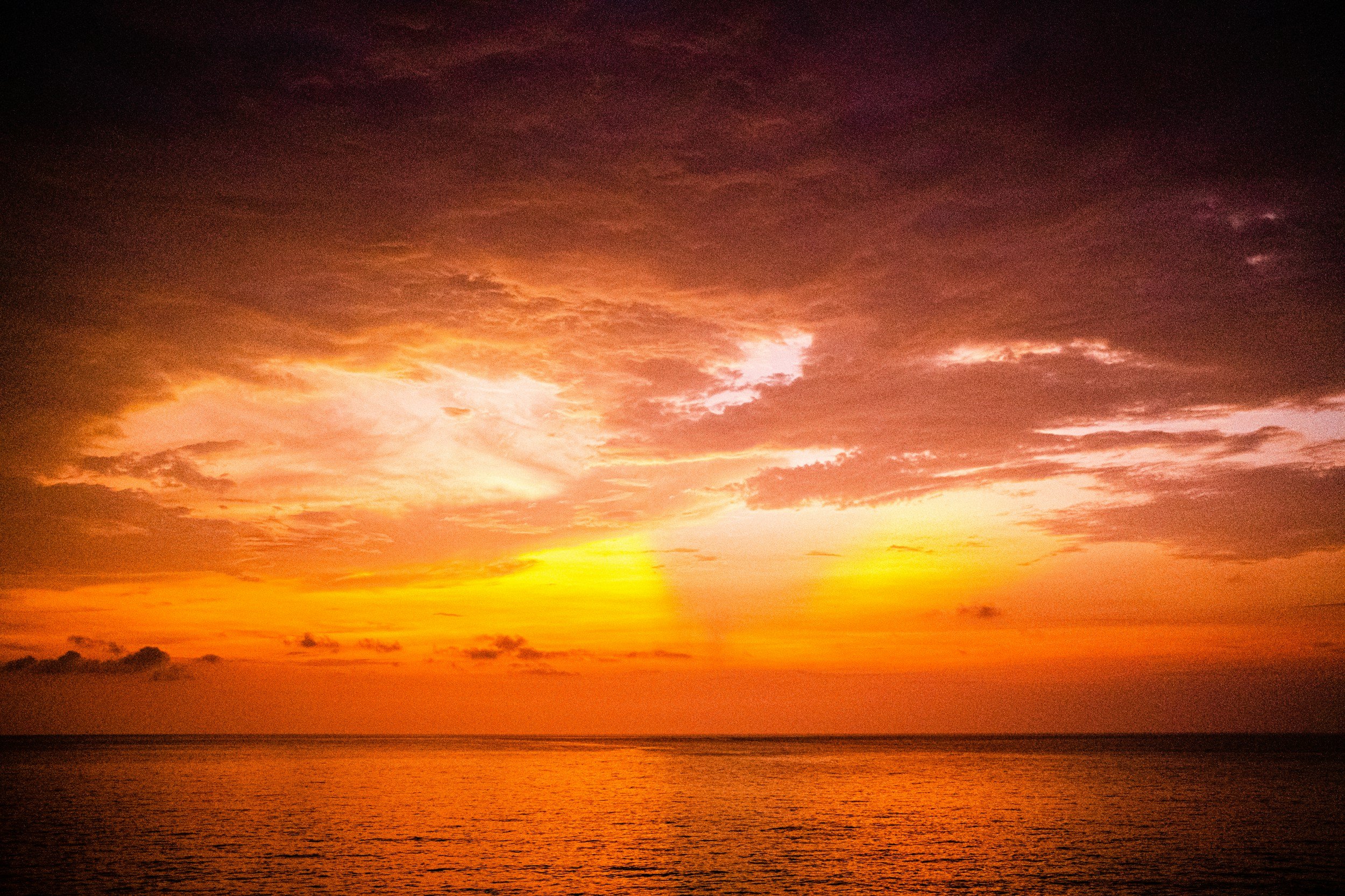 Sunset over the ocean with a sky filled with dark and orange clouds, reflecting orange and yellow hues on the water.