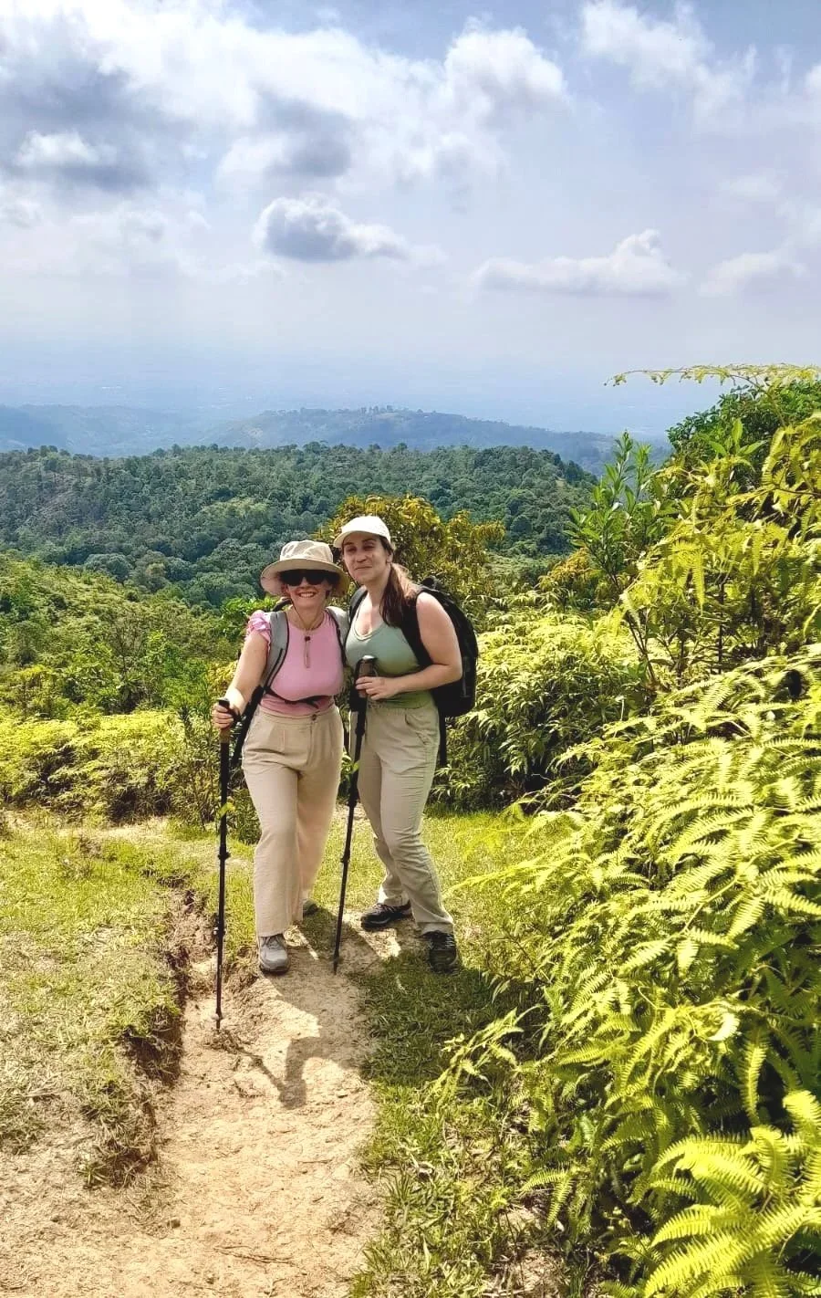 Two women on a hiking trail in a lush green mountain landscape under cloudy sky, carrying backpacks and walking sticks, smiling at the camera.