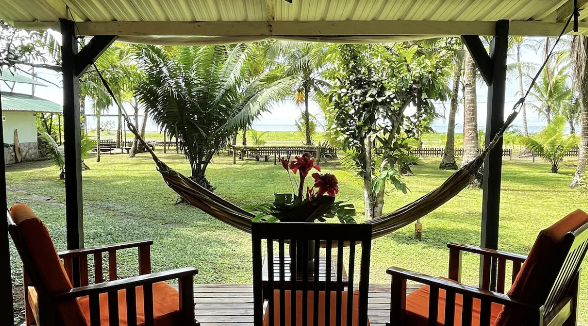 Porch view with hammock, chairs with cushions, and a table with a plant, overlooking a lush green tropical garden with palm trees and a glimpse of the ocean in the background.