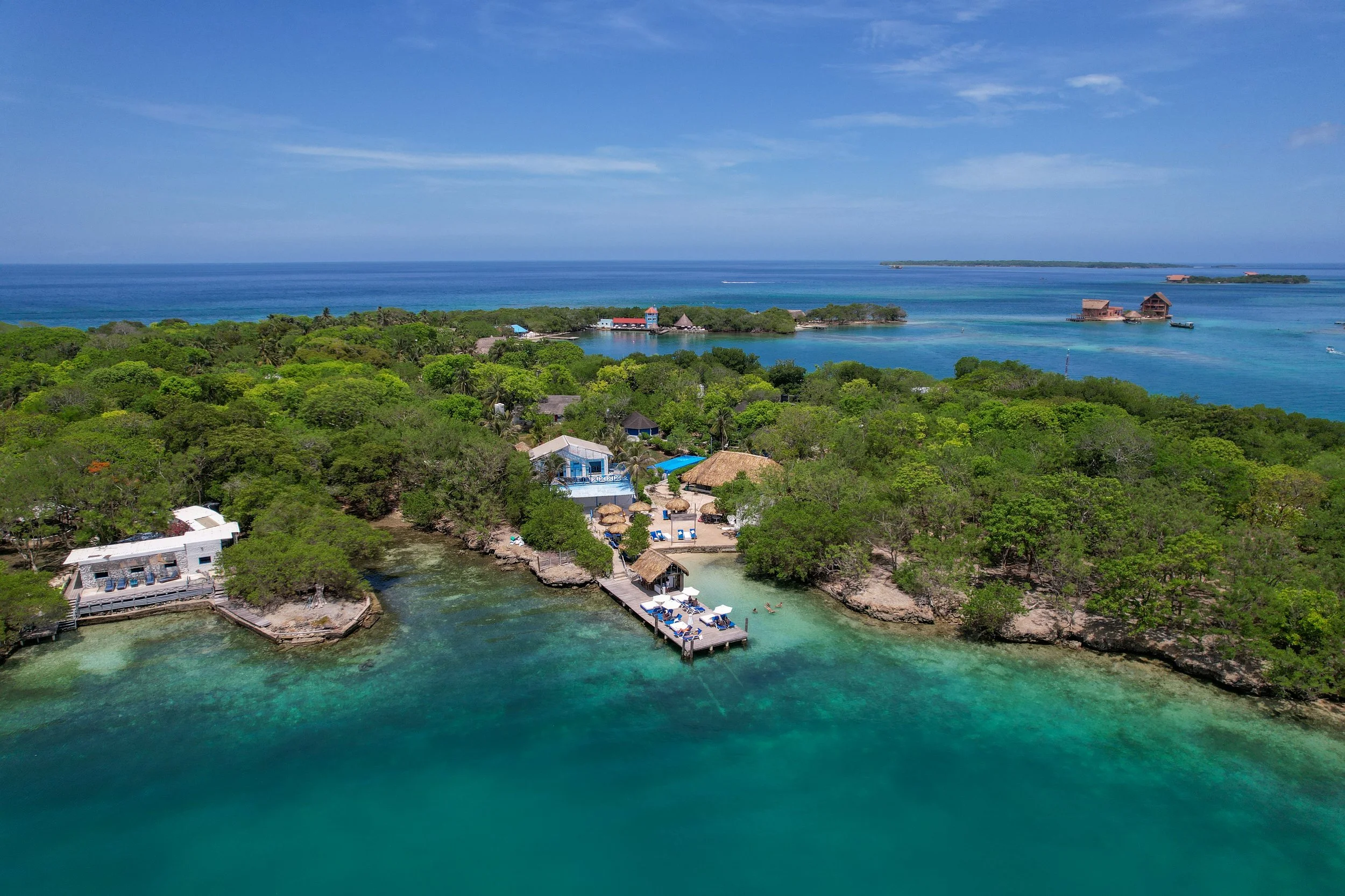 Aerial view of a tropical island resort with lush green trees, sandy beaches, and buildings extending into the turquoise ocean, with a clear blue sky overhead.