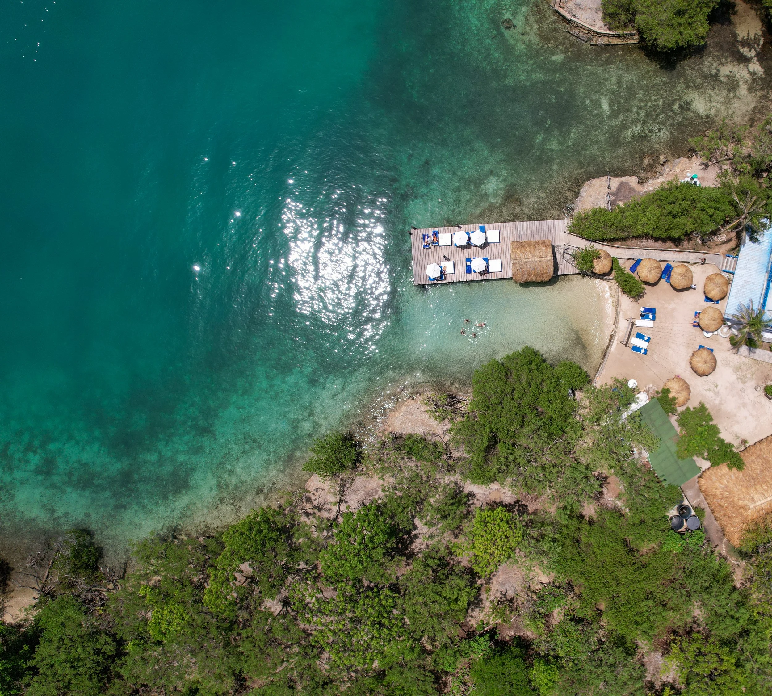 Aerial view of a beach with a wooden dock, sun umbrellas, lounge chairs, and trees along the shoreline in a coastal area.