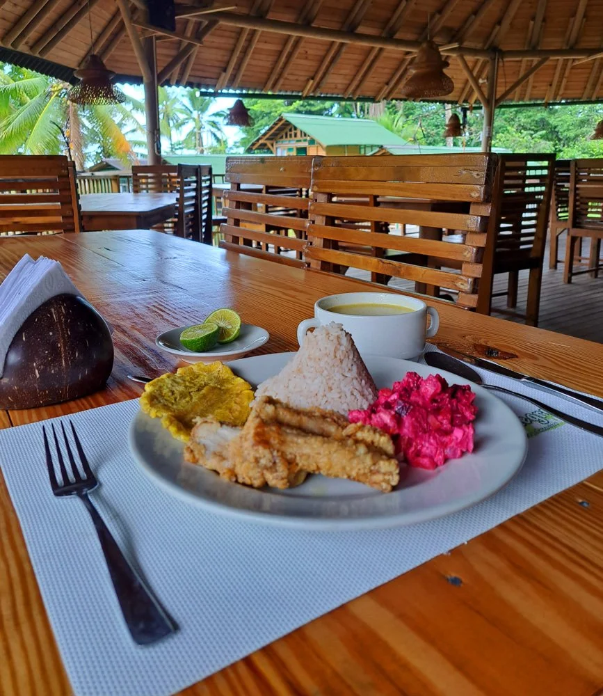 Plate of rice, fried chicken, pink coleslaw, fried plantain, and a cup of yellow soup or curry on a wooden table in an open-air restaurant with thatched roof and wooden furniture.