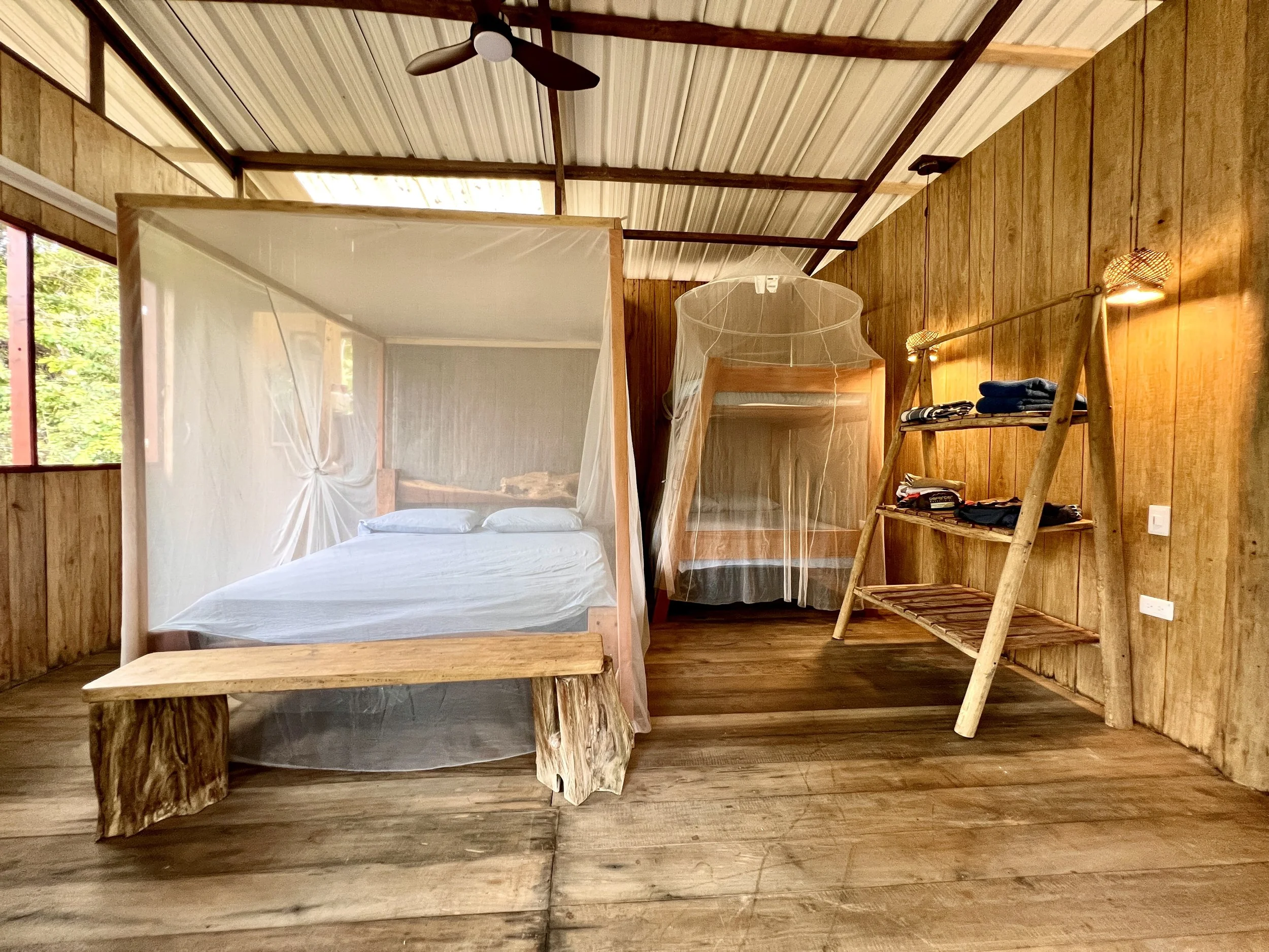A rustic bedroom with wooden walls and floor, featuring a large bed with white bedding and a mosquito net, a smaller bed with a mosquito net, a wooden ladder-style shelf with folded clothes, and a black ceiling fan.