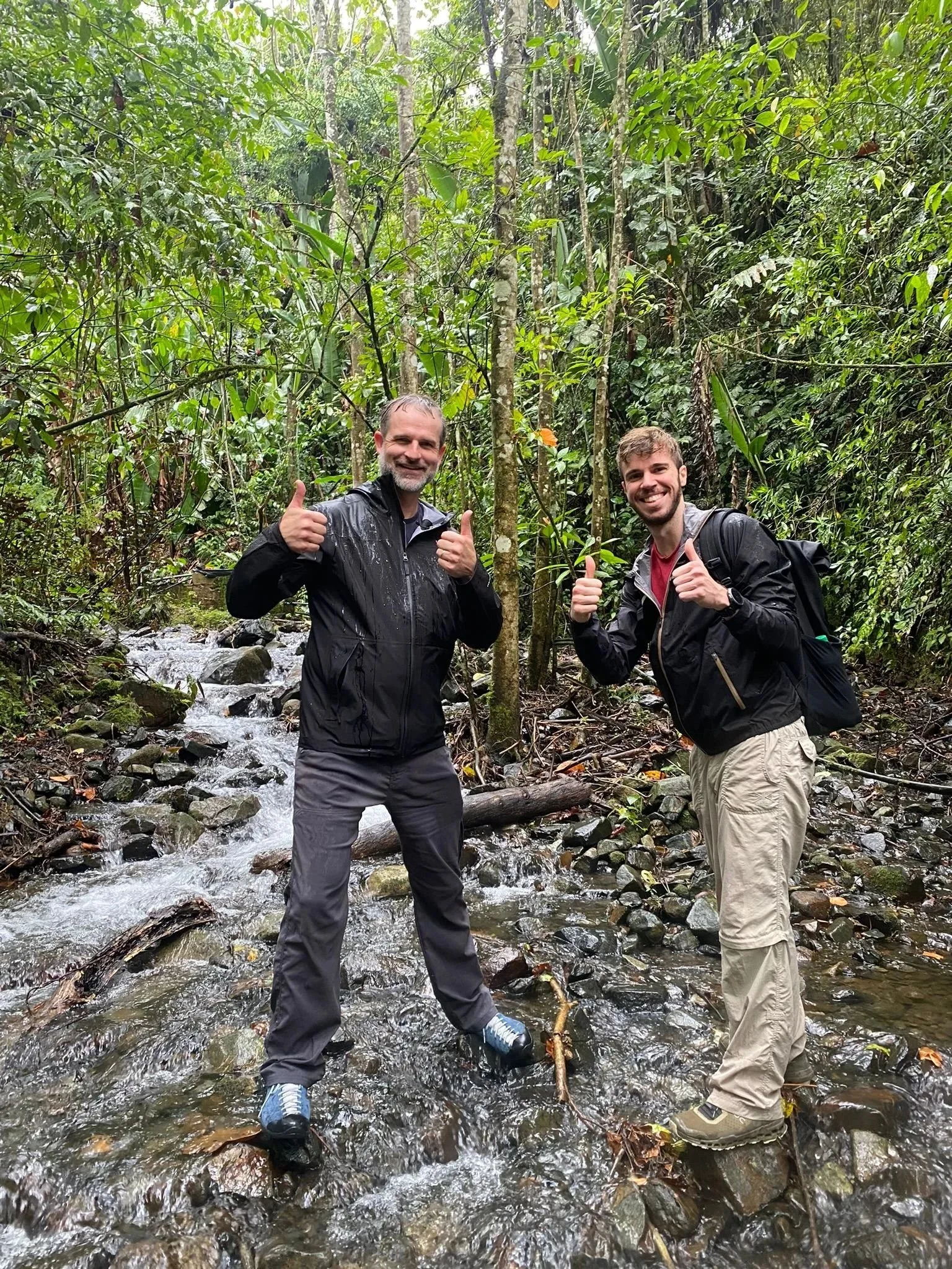 Two men standing in a shallow stream in a dense jungle, smiling and giving thumbs up, wearing waterproof jackets and hiking boots.
