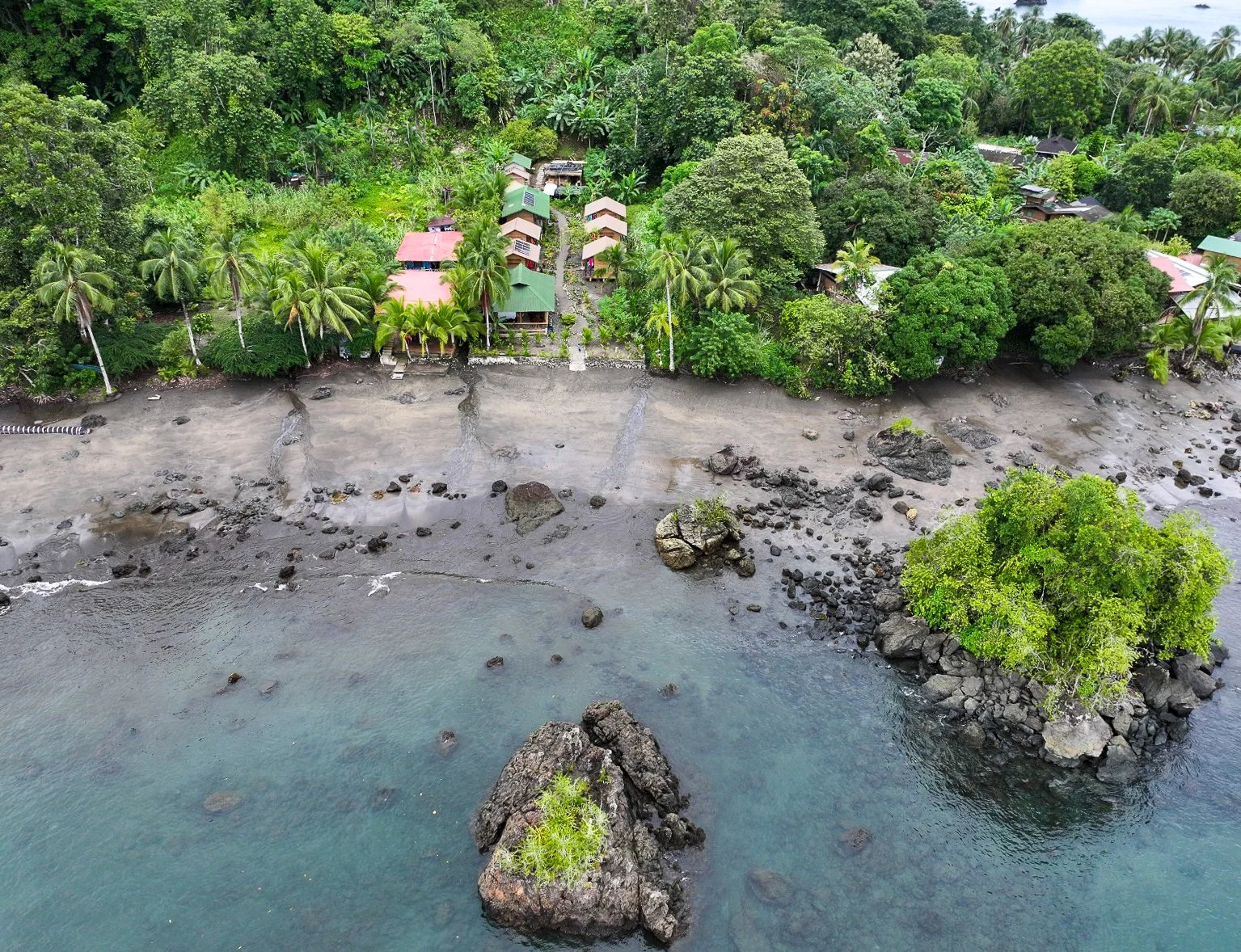 Aerial view of a tropical beach with rocks and clear water, lined with lush green trees and small houses with colorful roofs.