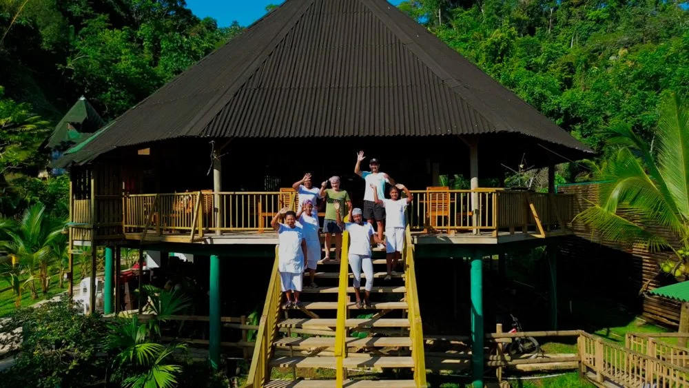 Group of six people standing on the stairs and porch of a stilt house with jungle surroundings.