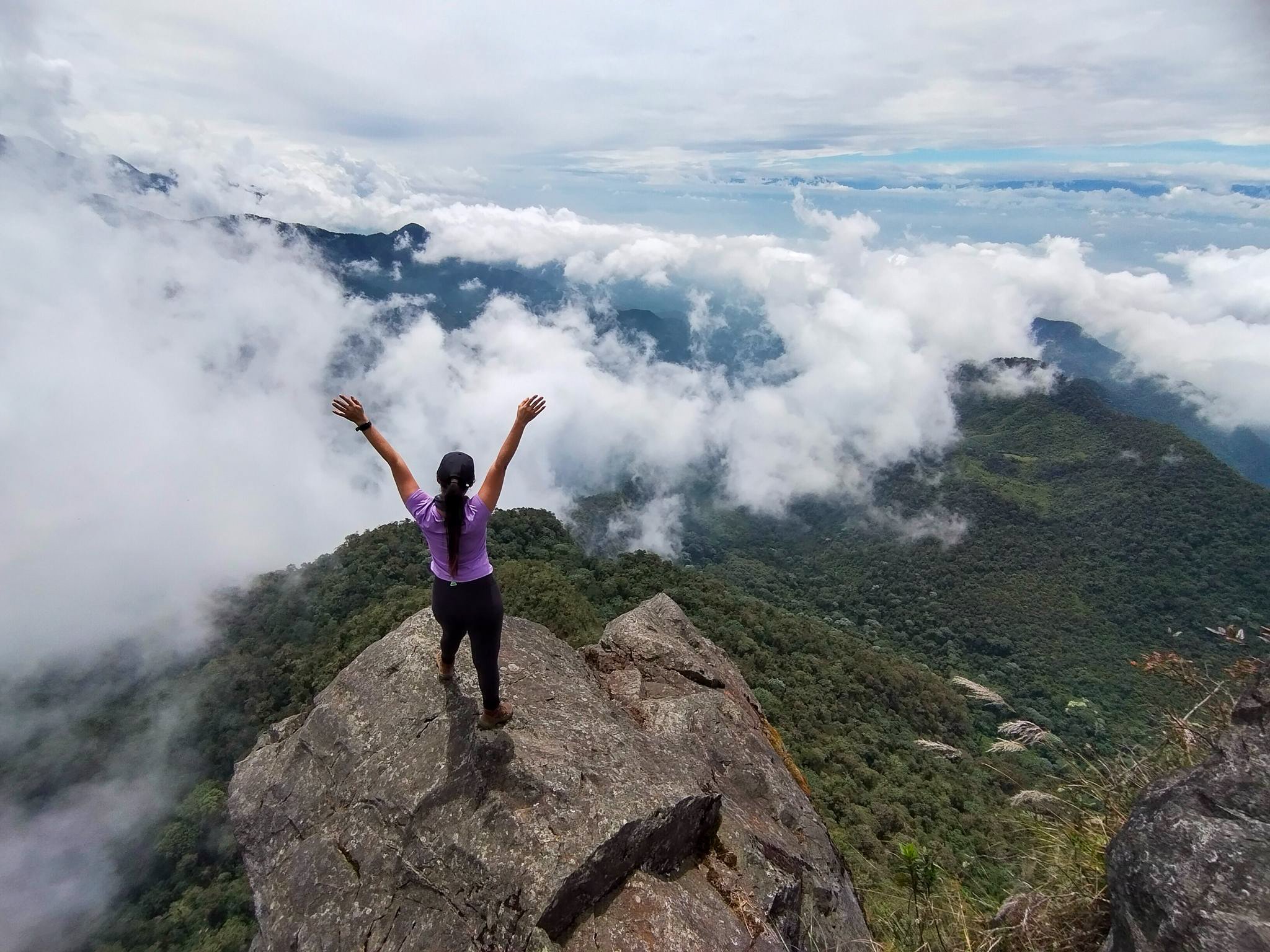 A person with arms raised standing on a rocky ledge on a mountain, looking at clouds and distant mountain peaks.