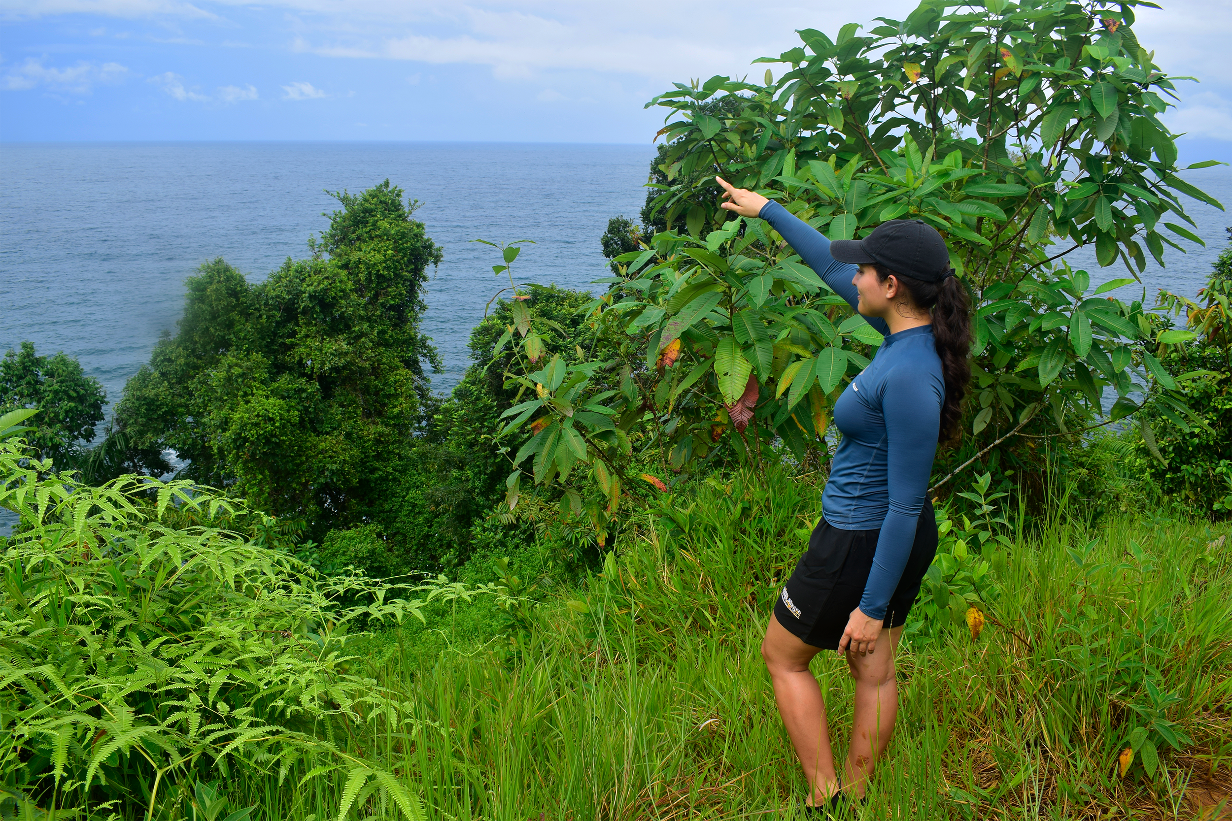 A woman in outdoor athletic clothing stands on a grassy ledge surrounded by lush green foliage, pointing towards the ocean visible in the distance under a partly cloudy sky.