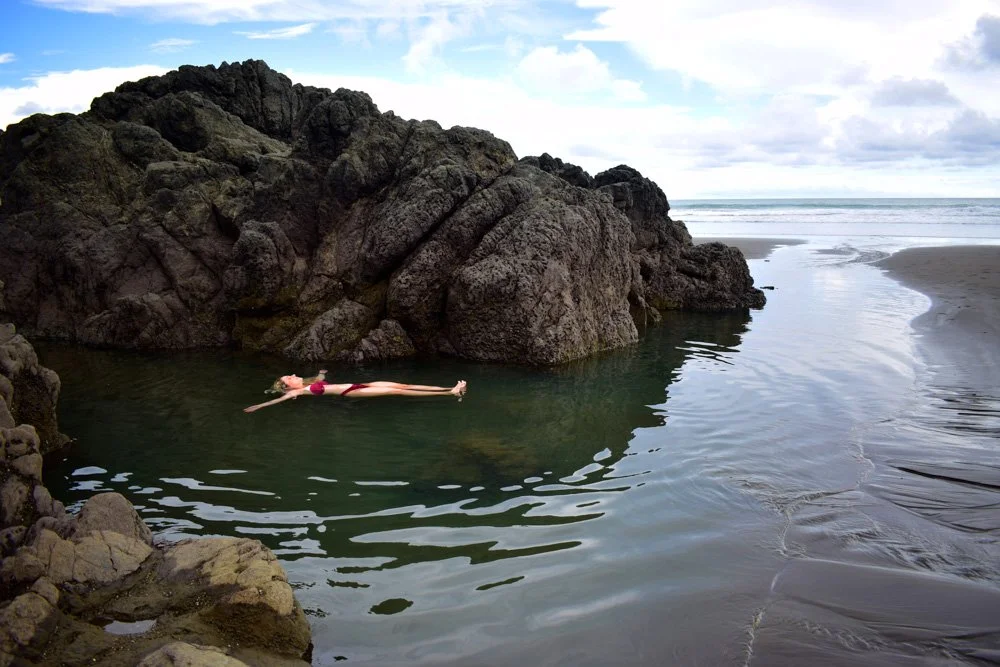 A woman in a red swimsuit floating on her back in a small pool of water formed by rocks on a beach, with the ocean and cloudy sky in the background.