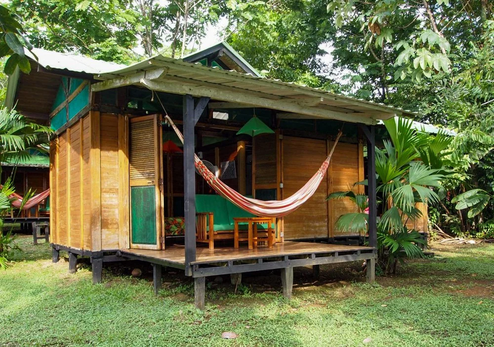 A small wooden cabin with an open porch, hammock, table, and lush green plants surrounding it in a tropical setting.