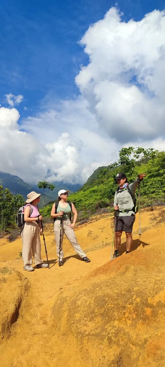 Three hikers with backpacks and trekking poles stand on a yellow dirt trail in a mountainous area with lush green vegetation and cloudy sky.