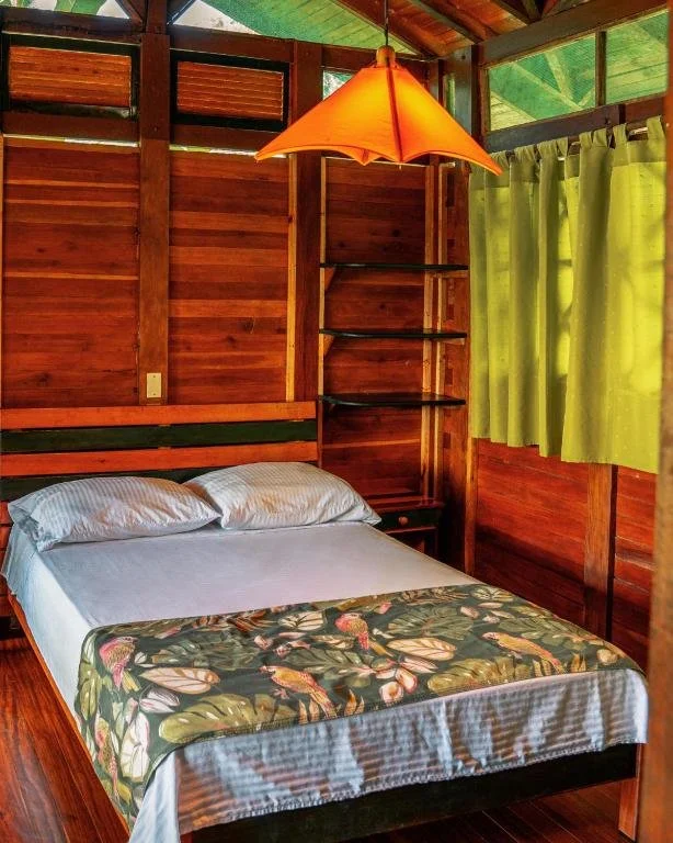 Wood-paneled bedroom with a bed, white pillows, a floral bedspread, a hanging orange lampshade, a window with green curtains, and built-in shelves.