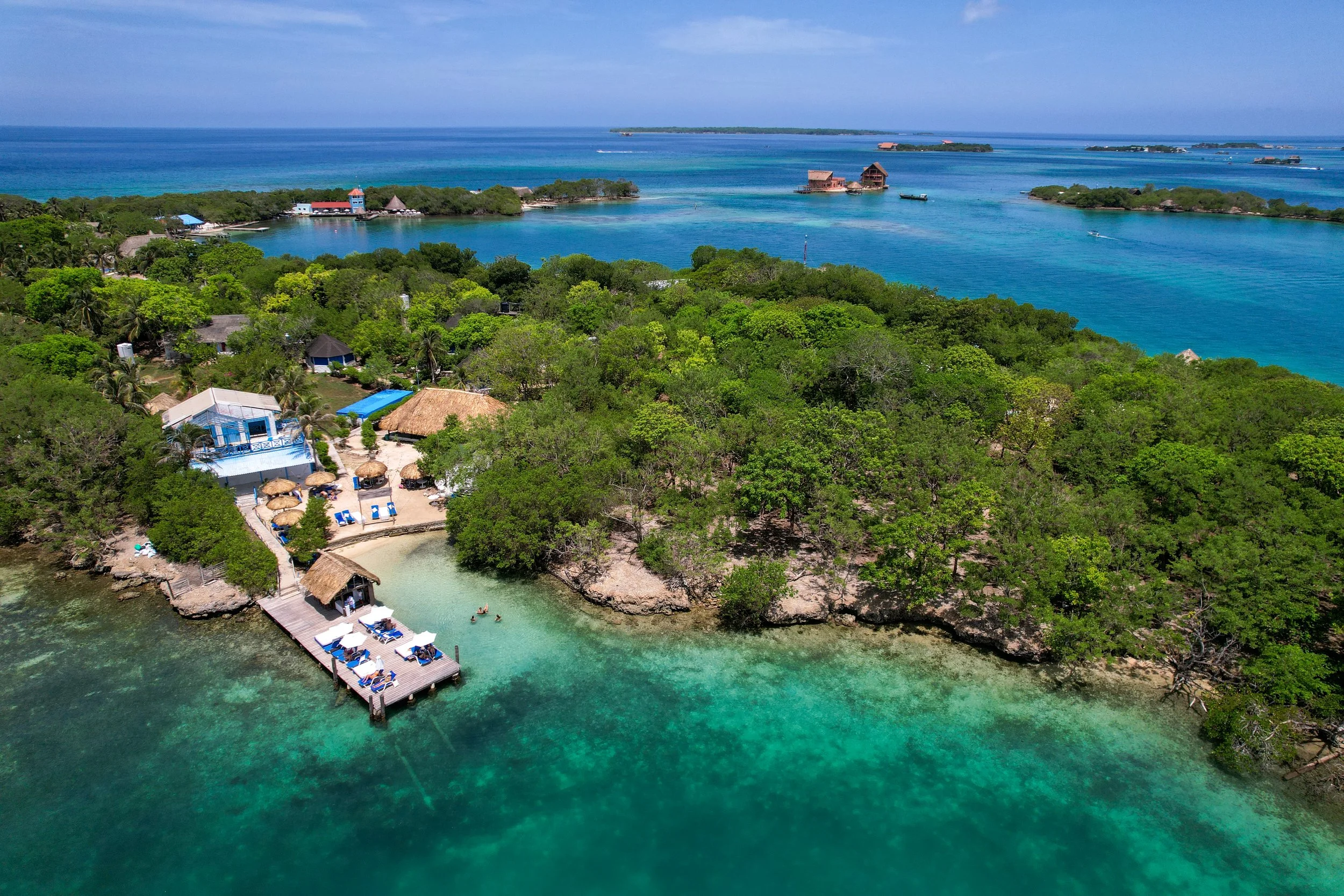 Aerial view of a tropical resort with a beach, lush green trees, and clear turquoise water, including a pier with lounge chairs, some swimmers, and buildings surrounded by dense vegetation.