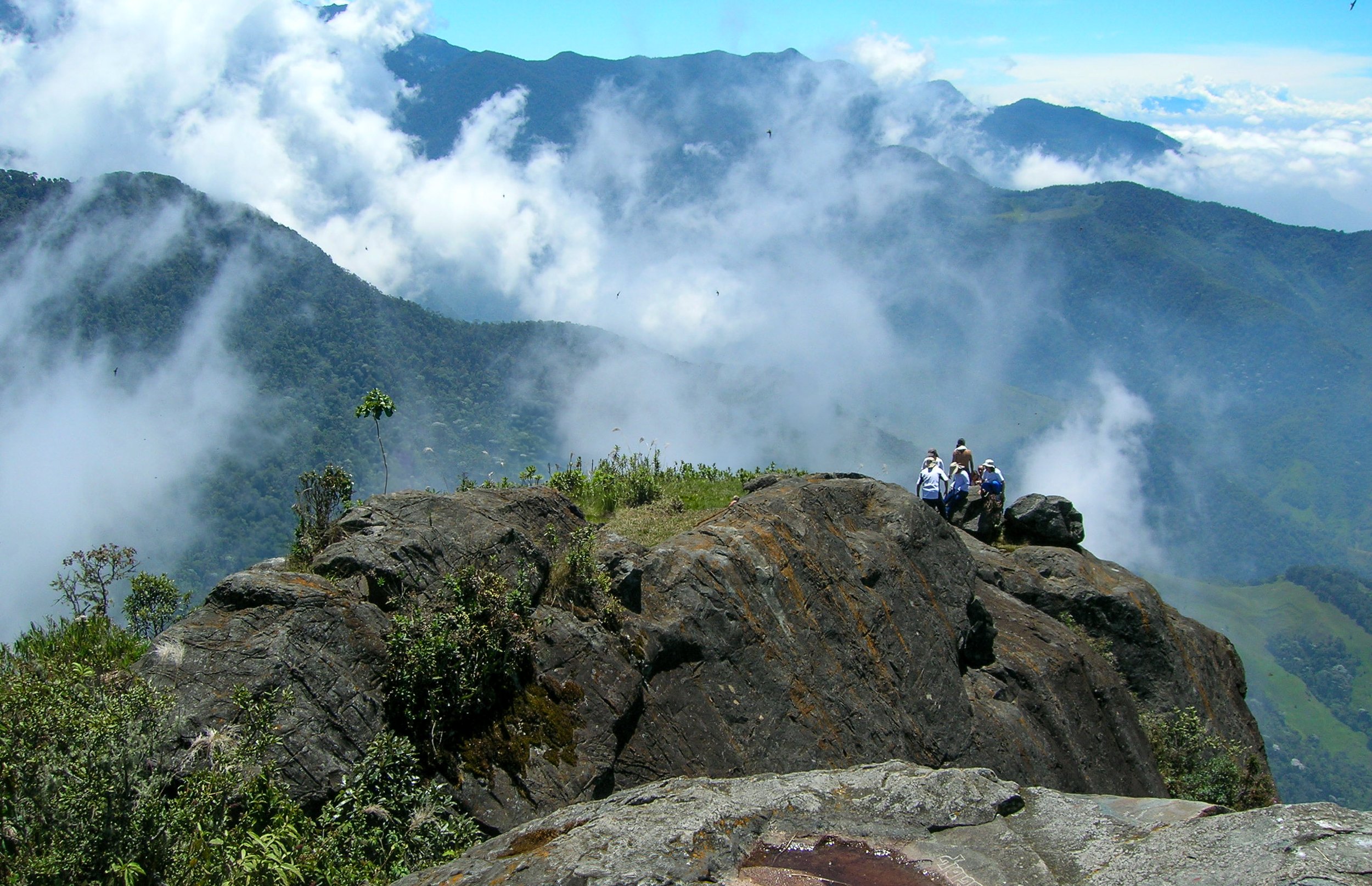 View of mountains with clouds and a group of four people sitting on rocks in the foreground.