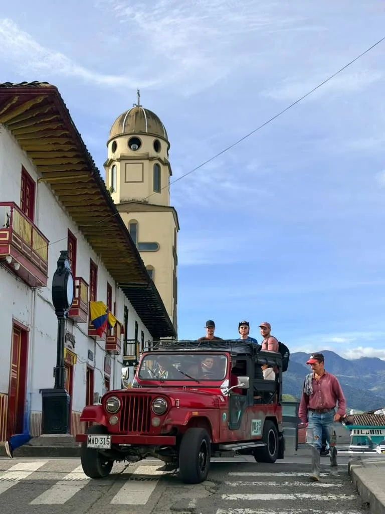 Tourists riding in a red open-top jeep on a city street with colonial-style buildings, a clock tower, and mountains in the background.