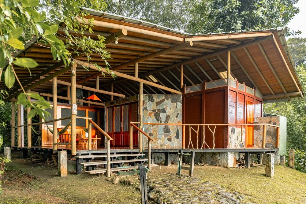 Rustic cabin with wooden and stone exterior, elevated on stilts, surrounded by green trees, with an open front porch featuring wooden furniture and a stone path leading up to it.