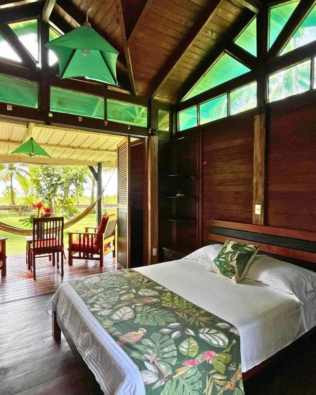 A cozy bedroom with wooden walls and a large bed decorated with tropical leaf-themed bedspread and pillows. There is a small dark wood shelf in the corner. The room opens to a porch with two chairs and a hammock, overlooking a lush green landscape wi
