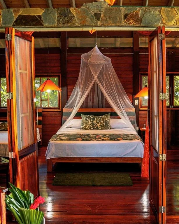 A cozy bedroom with a wooden four-poster bed featuring a mosquito net, tropical-themed bedding, and decorative pillows. The room has wooden walls, small windows with natural light, and warm lighting from bedside lamps.