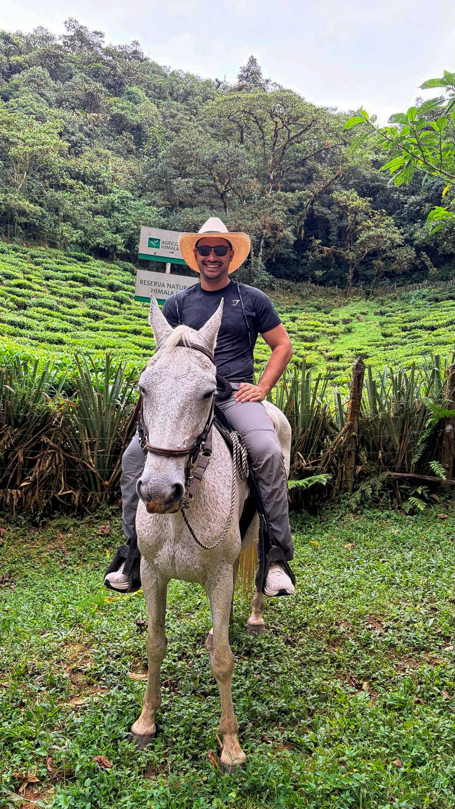 Man riding a white horse in a lush, green landscape with trees, grass, and a sign that reads 'Reserva Natural Himalaya' and 'Agricultores Himalaya'.
