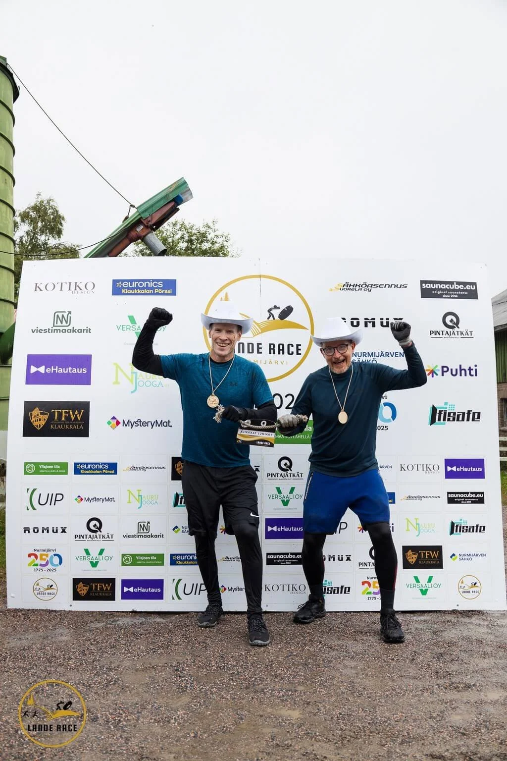 Lande Race Nurmijärvi on extremerun-tyylinen OCR-hupijuoksu. Kuvassa kaksi osallistujaa juhlii cowboy-hatuissa ja mitaleissa sponsoriseinän edessä.