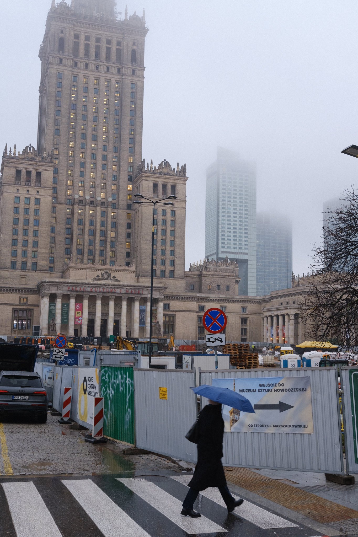 Street photo of Warsaw showing a person with an umbrella walking in front of the Palace of Culture and Science.