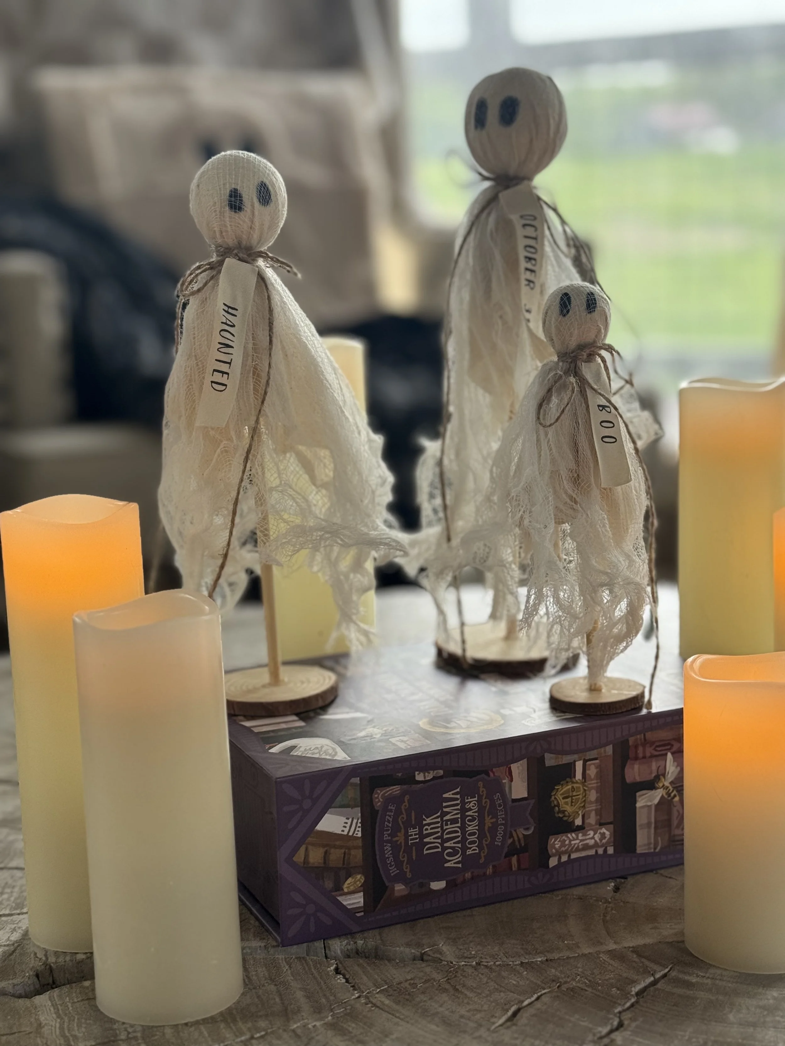 Decorative figurines resembling ghostly characters with fabric and string features, labeled 'Haunted' and 'October,' surrounded by candles and a box of a puzzle titled 'The Dark Academic Bookcase' on a rustic wooden surface.
