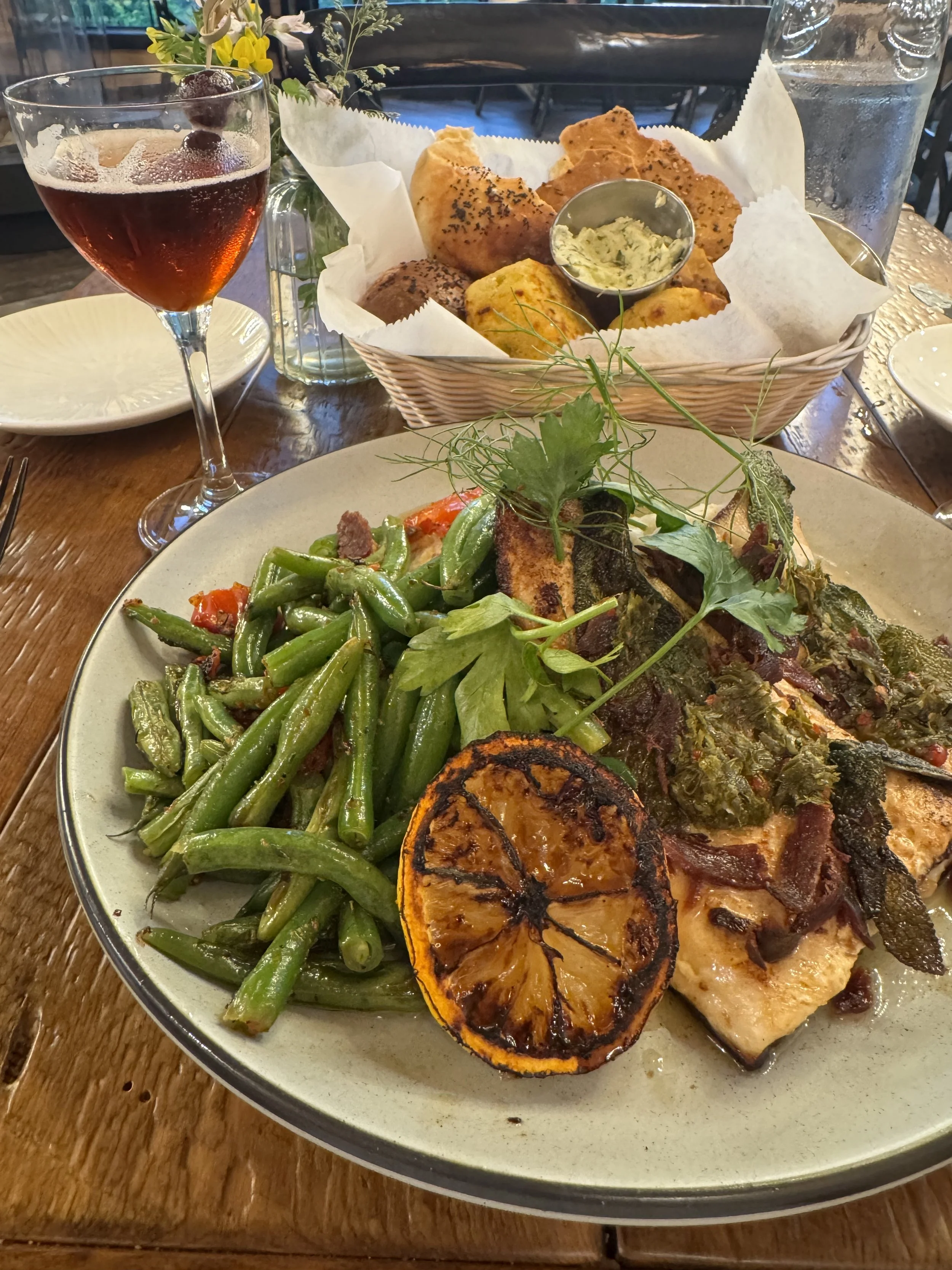 A plate of grilled vegetables, including grilled zucchini slices, green beans, and leafy greens, with a garnish of fresh herbs. Behind the plate, there is a basket of fried foods with a small container of dip, a glass of red drink with berries, and a large glass of water.