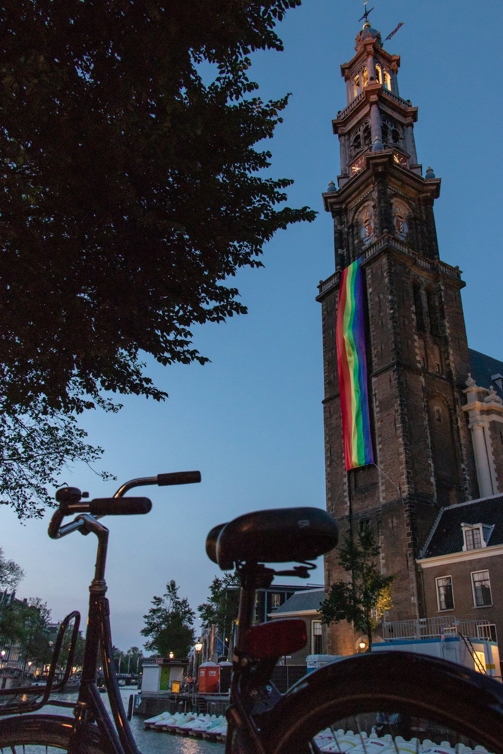 A tall brick church tower with a rainbow pride flag hanging from it, trees, and bicycles in the foreground, during twilight.