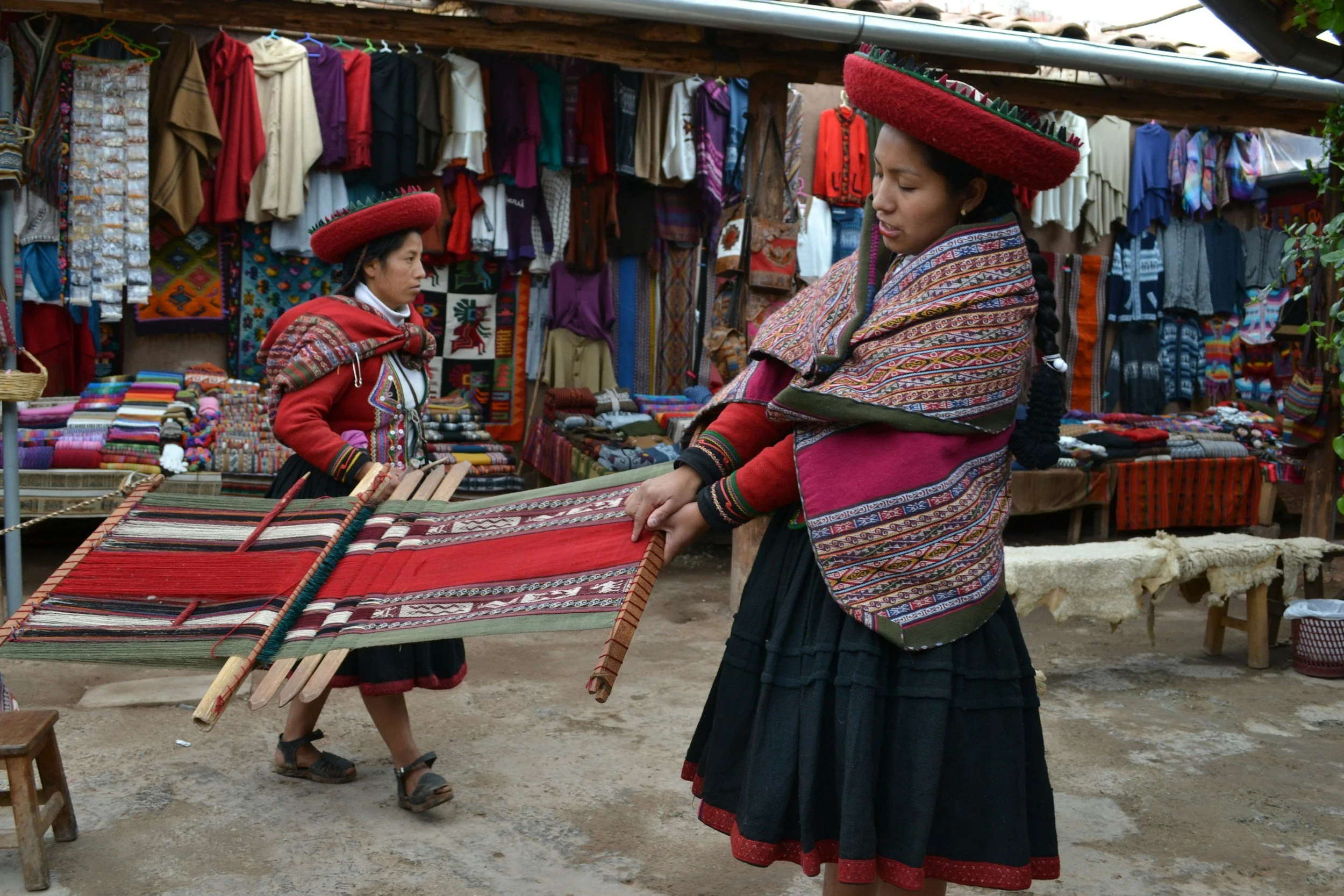 Two women dressed in traditional Andean clothing at a marketplace, with colorful textiles and garments hanging in the background. One woman is holding a woven cloth, and both women are wearing large, circular hats.