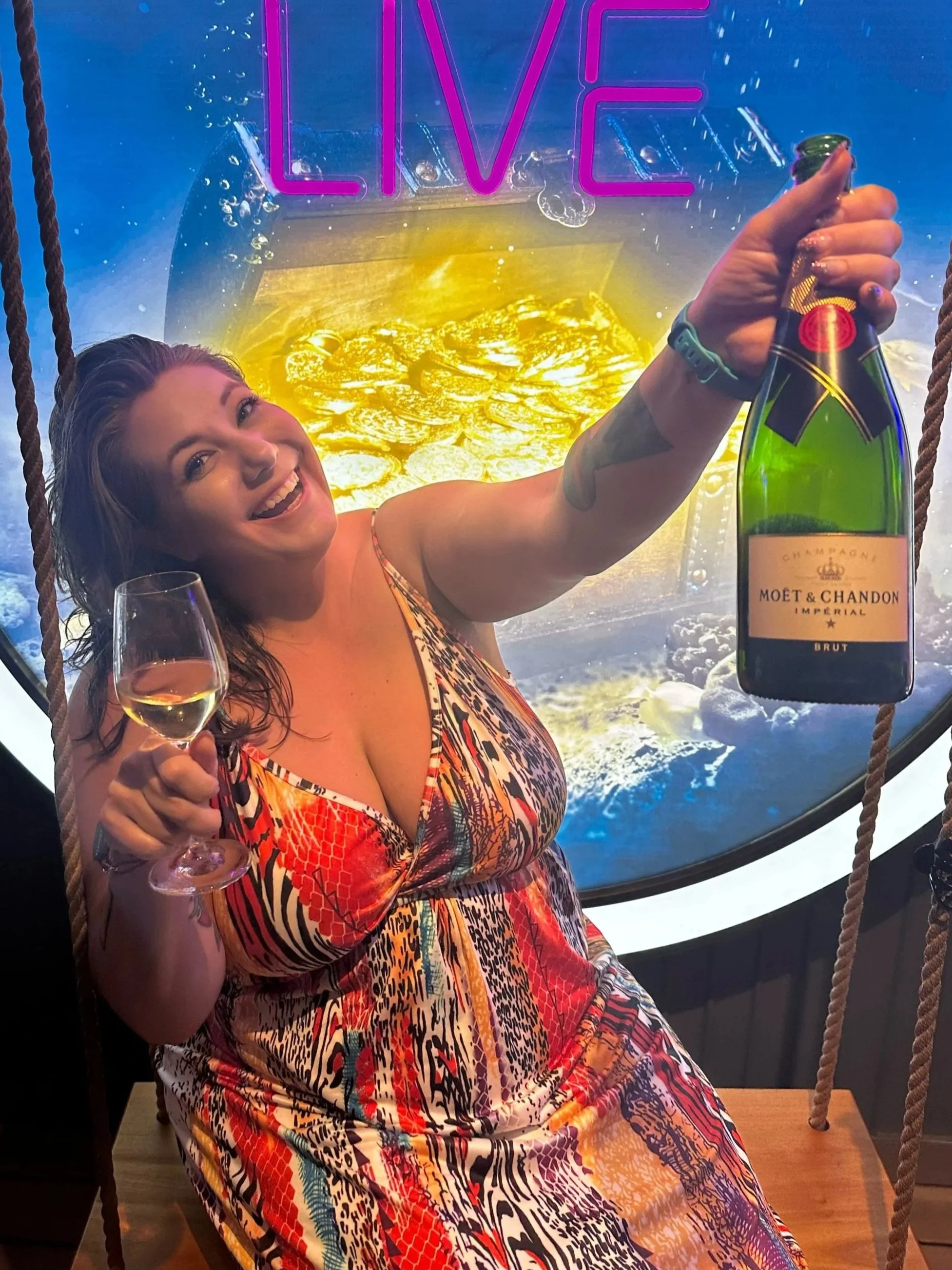 A woman smiling and holding a glass of wine in one hand and a champagne bottle in the other, sitting in front of a neon sign that says 'LIVE' with a background of a large gold-colored object, possibly a treasure chest filled with gold coins.