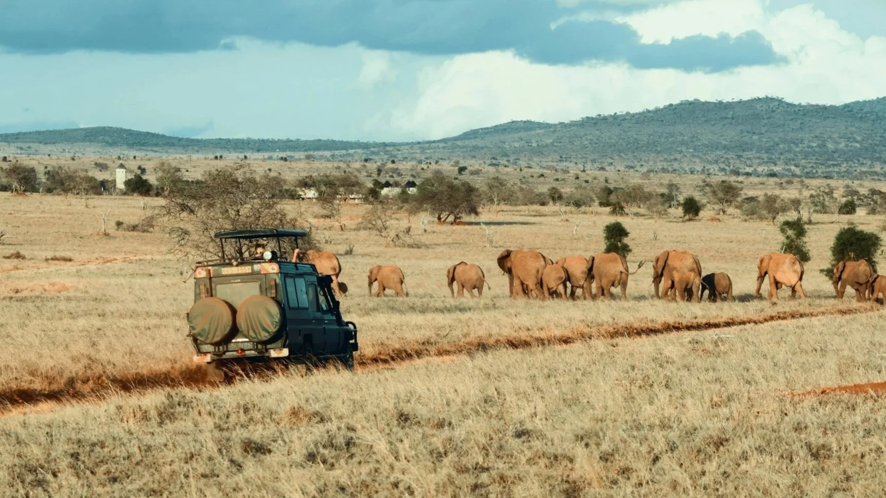A land vehicle driving on a trail in an African safari landscape, with herd of elephants in the background, some trees, and a mountain range under a partly cloudy sky.