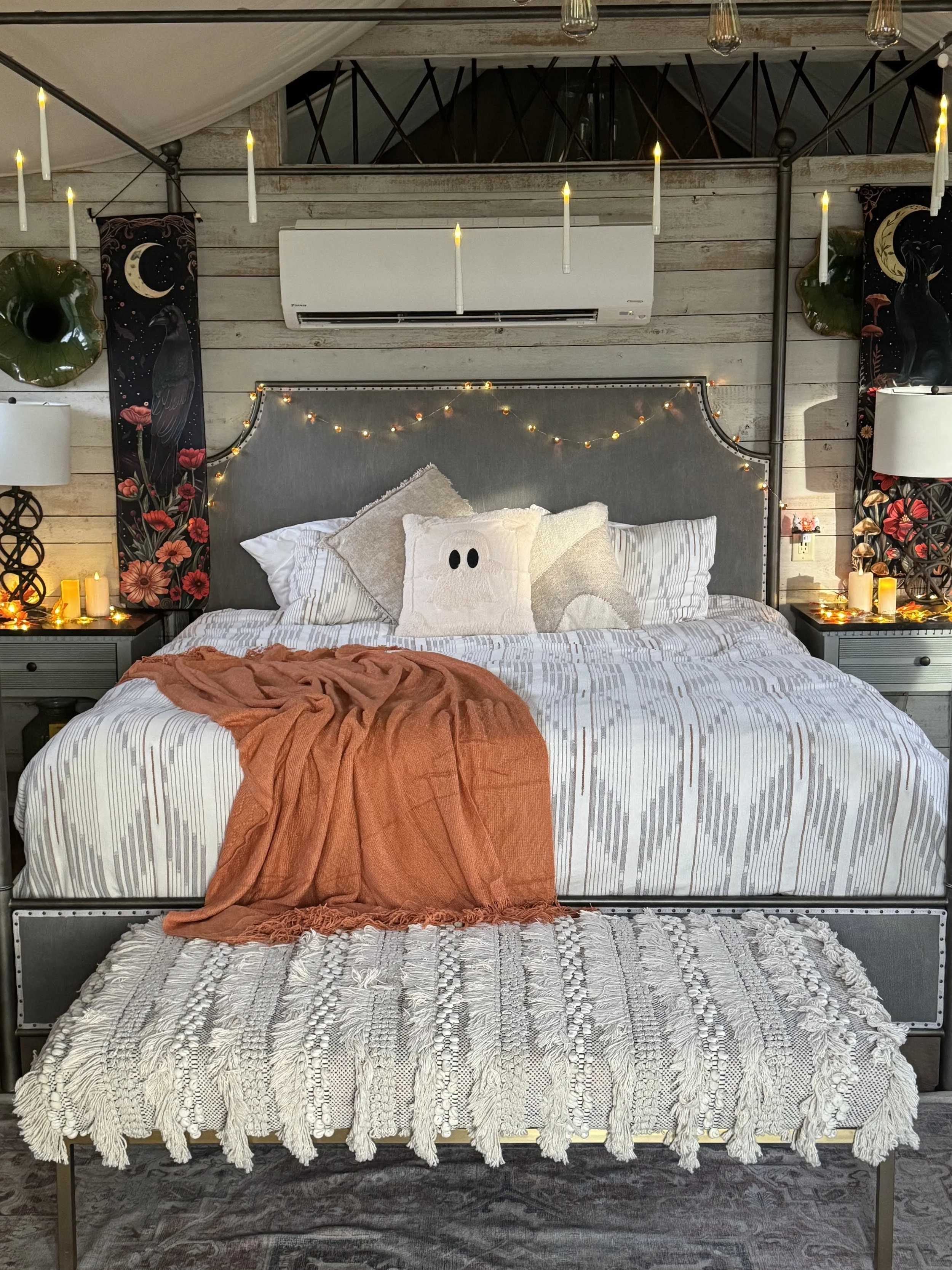 A neatly made bed with white striped bedding, decorated with a beige pillow and a plush ghost pillow, topped with a burnt orange blanket. The headboard has fairy lights. Two nightstands with candles and lamps flank the bed. The wall behind features wooden panels with hanging art, candles, and an air conditioning unit.