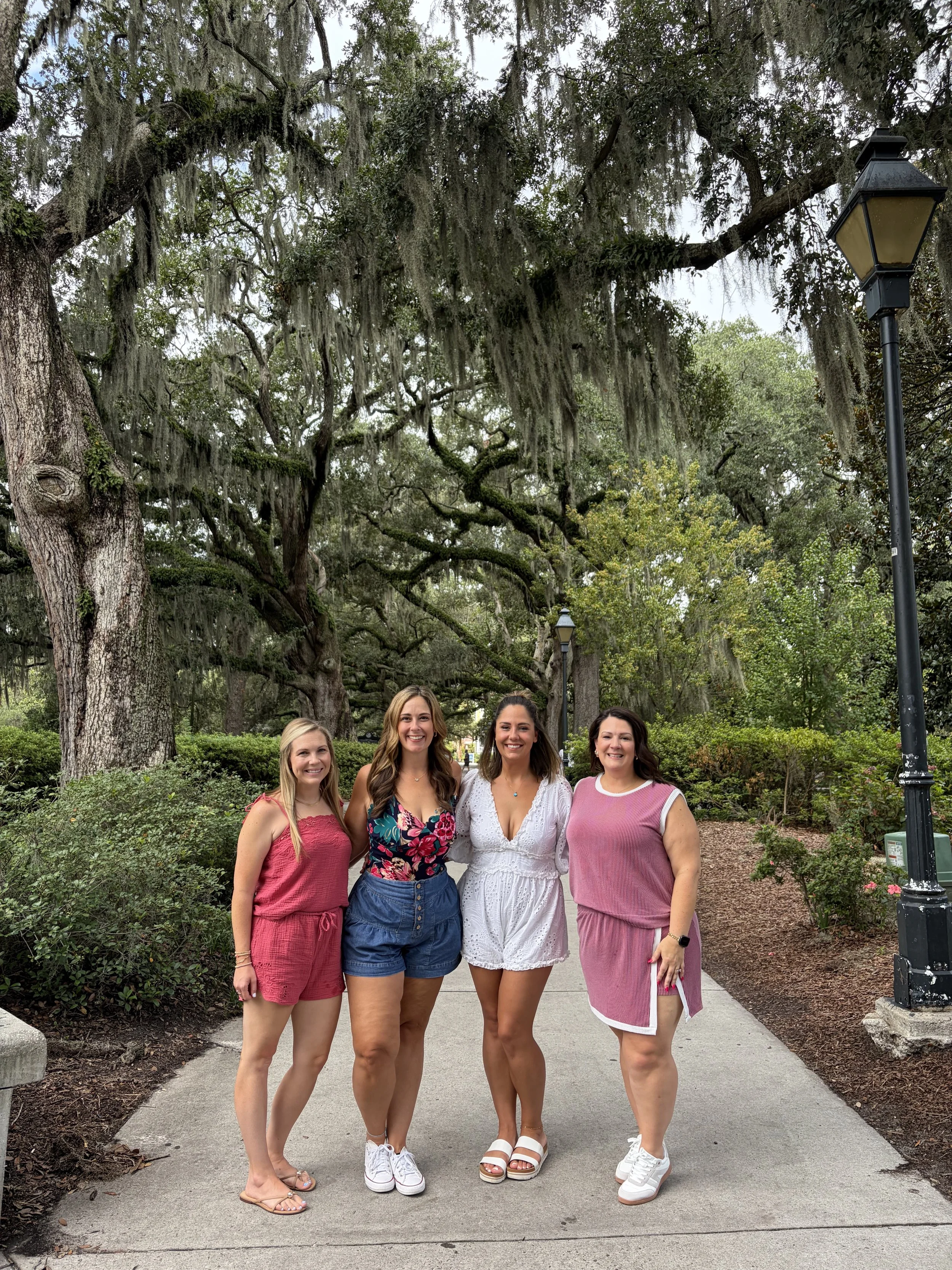 Group of four women smiling together on a paved walkway under mossy oak trees in Savannah Georgia.
