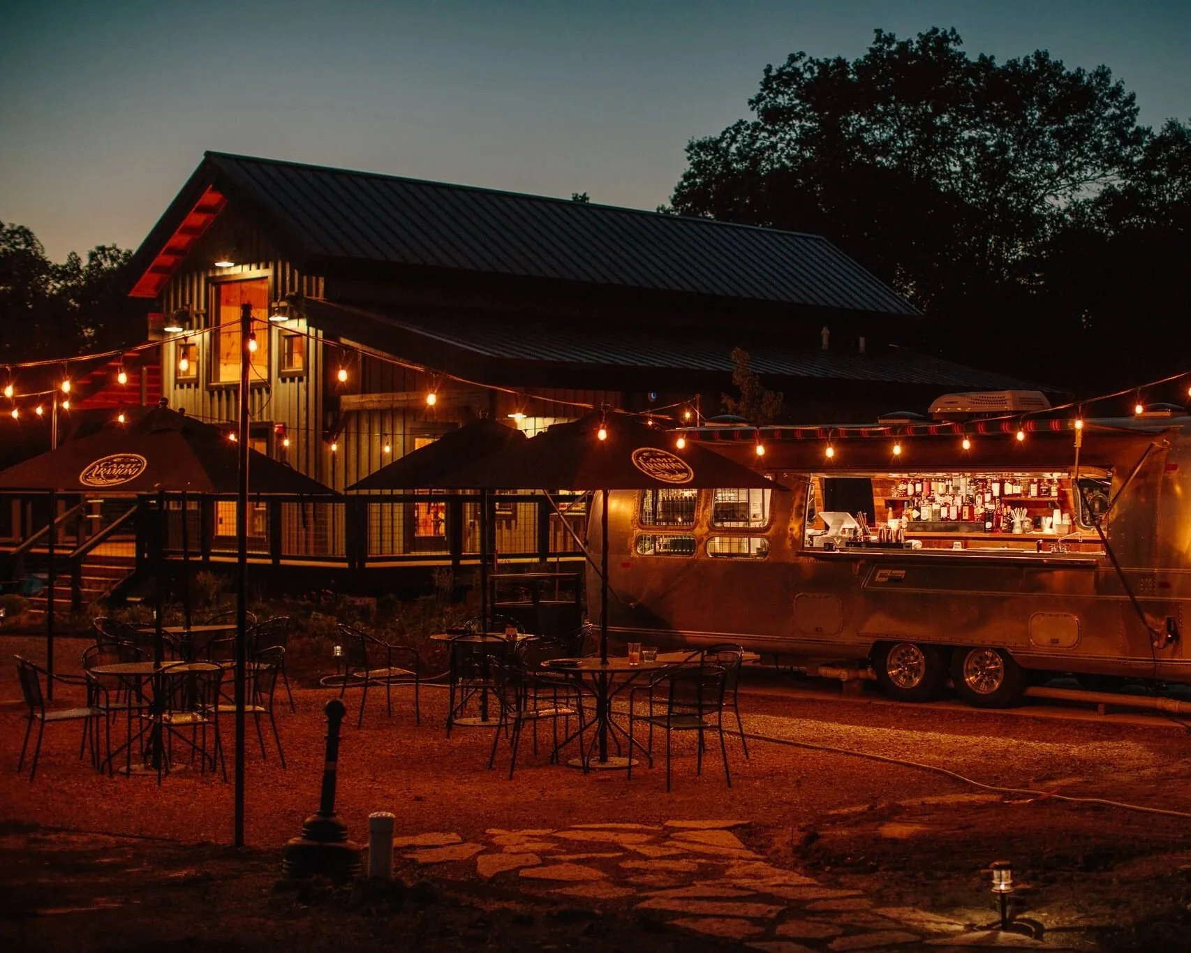A nighttime scene of an outdoor dining area illuminated by string lights, with a food truck serving drinks and decorations, next to a building with a metal roof surrounded by trees.