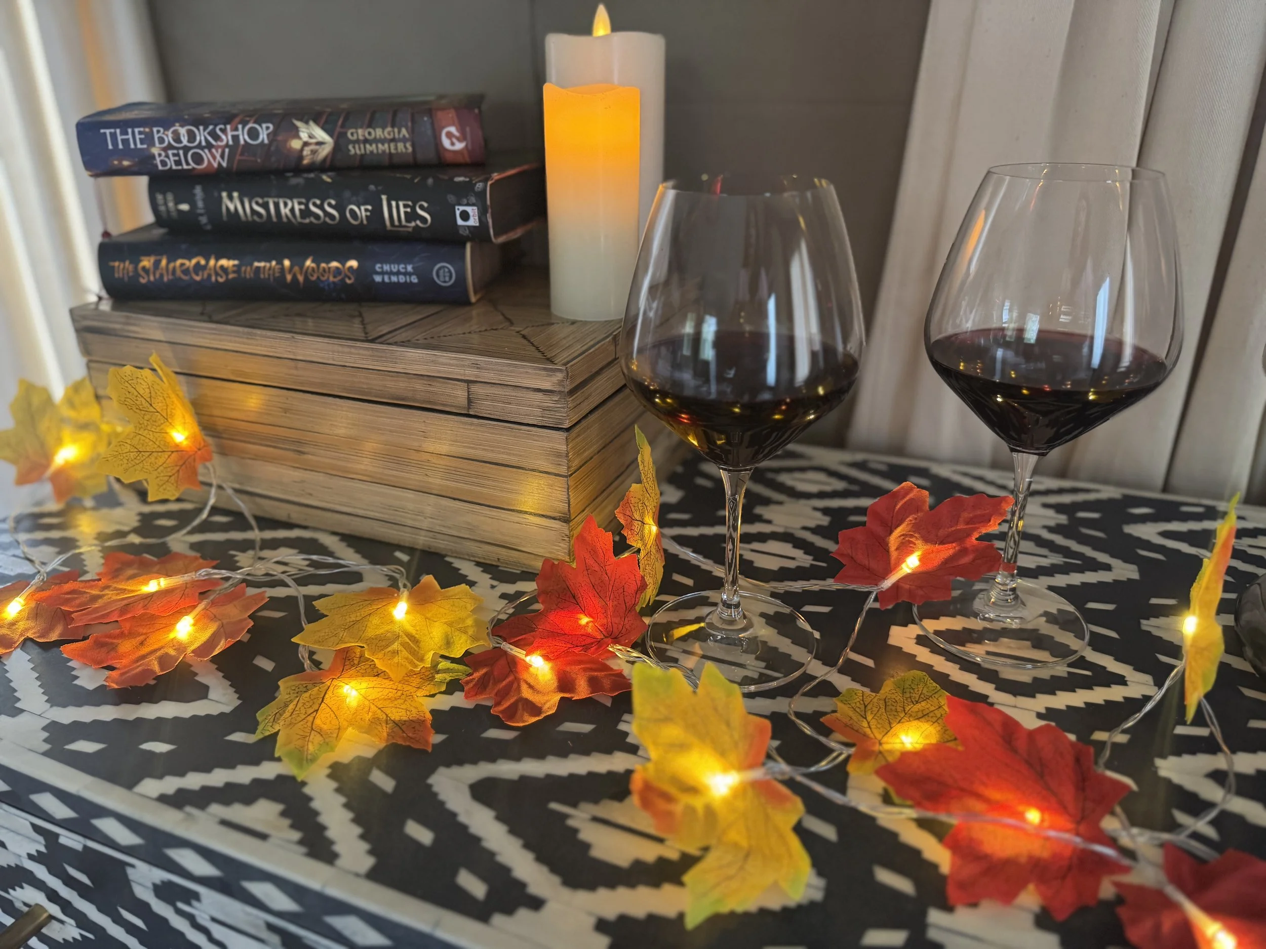 A table decorated with a string of autumn leaves and small lights, with two glasses of red wine, a stack of three books, a lit candle, and a white candle in the background.