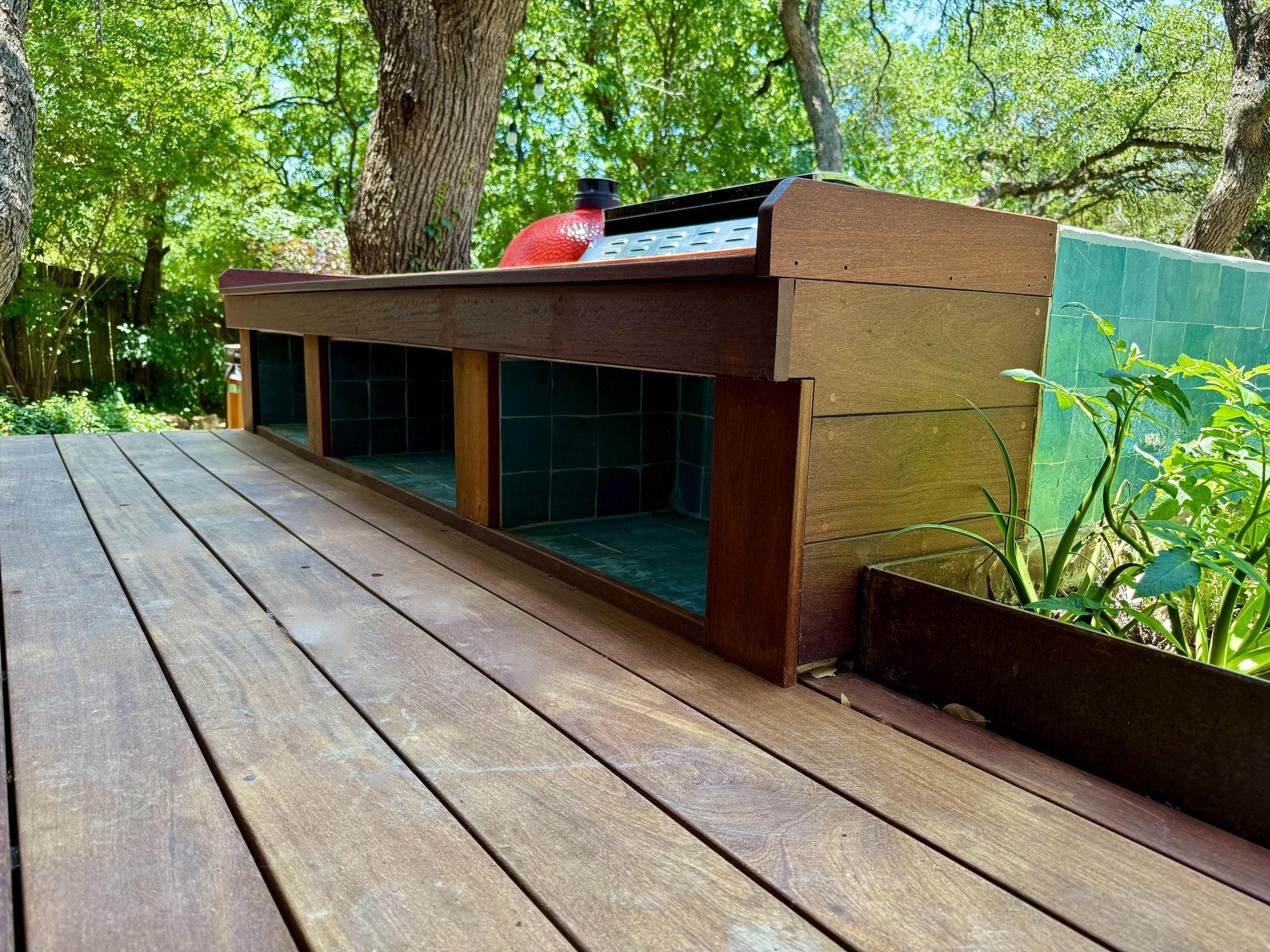 Outdoor wooden deck with a built-in barbecue grill surrounded by trees and greenery.