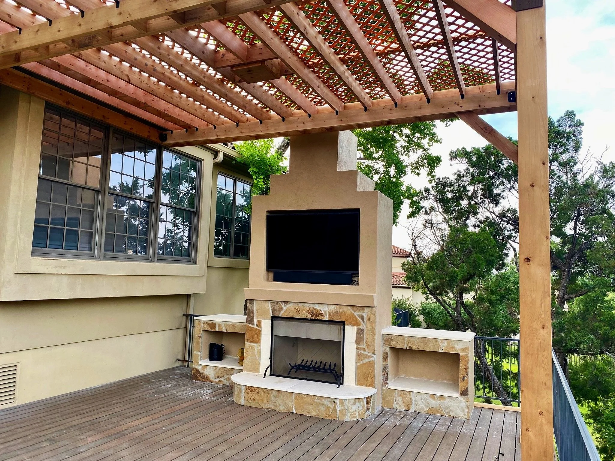 Outdoor patio with a stone fireplace, mounted flat-screen TV, wooden deck flooring, and lattice-covered wooden pergola, with large windows and trees in the background.