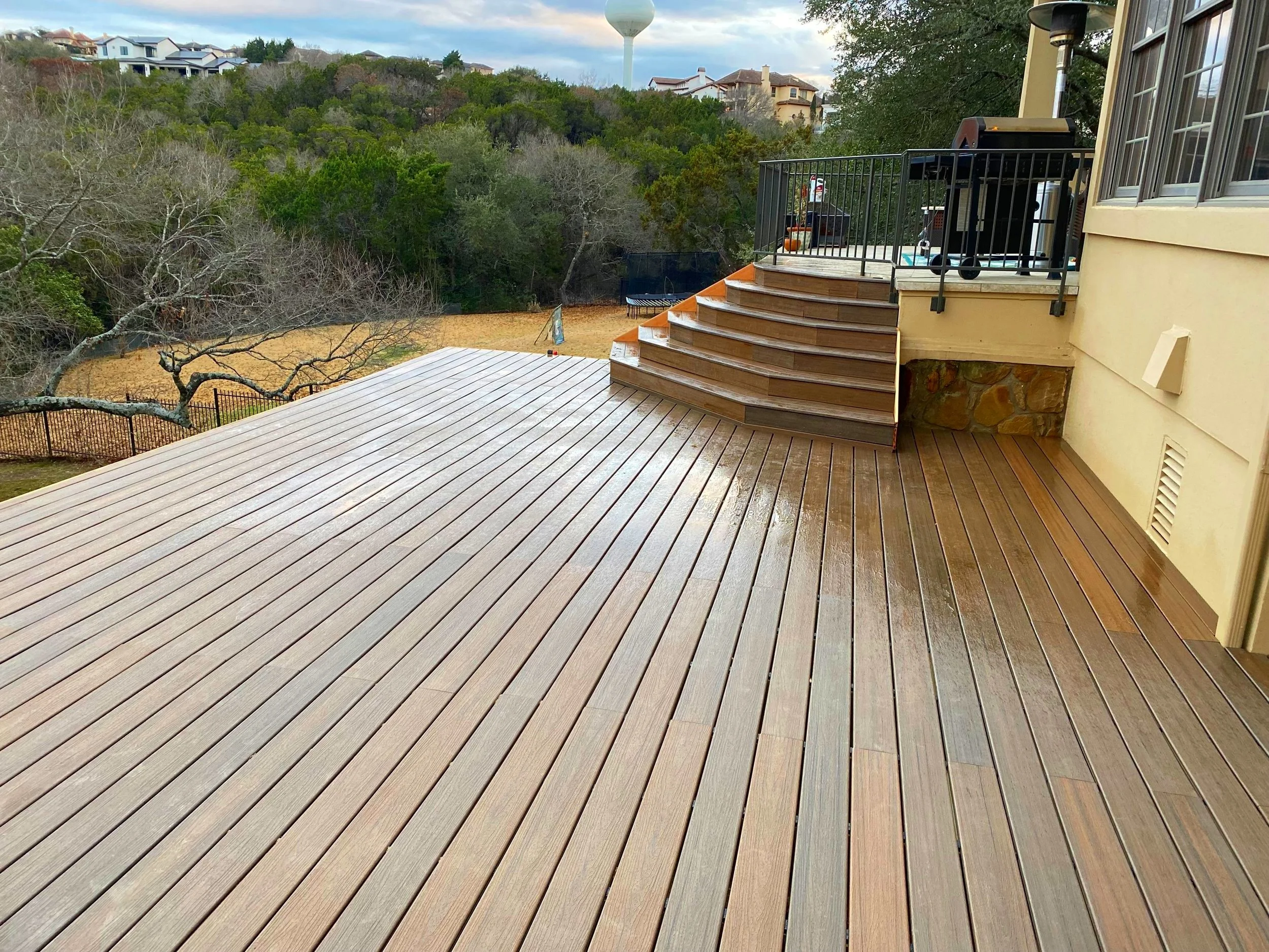 Clean, newly finished wooden deck with steps leading to elevated balcony, overlooking a backyard with trees and houses in the distance.
